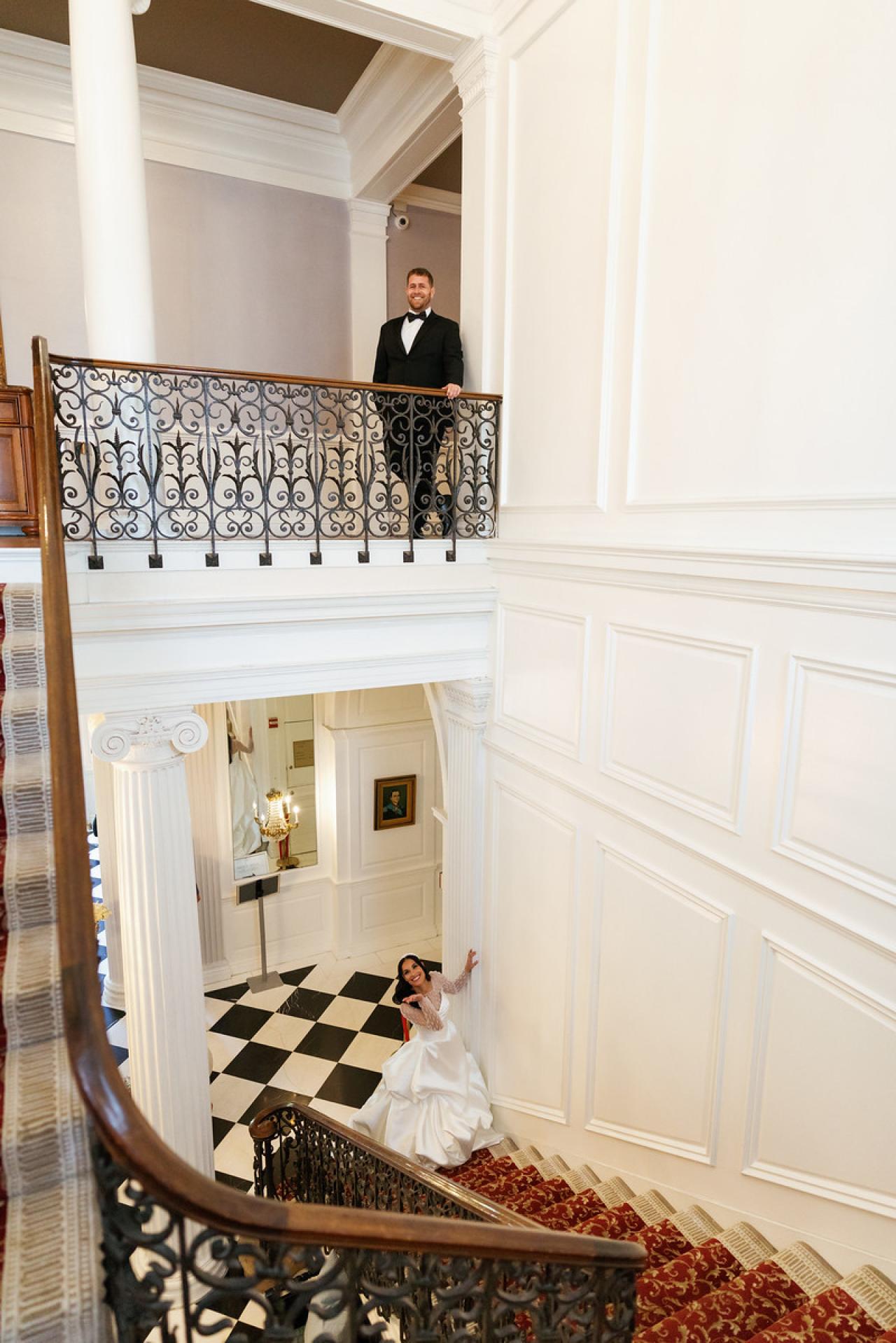 Bride standing in a white dress on a lower level while the groom in at The Mansion At Glen Cove, photographed by CJ Studios. Wedding photography.