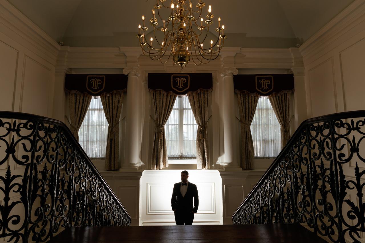 Silhouetted groom in a black suit and bow tie standing on a grand at The Mansion At Glen Cove, photographed by CJ Studios. Wedding photography.