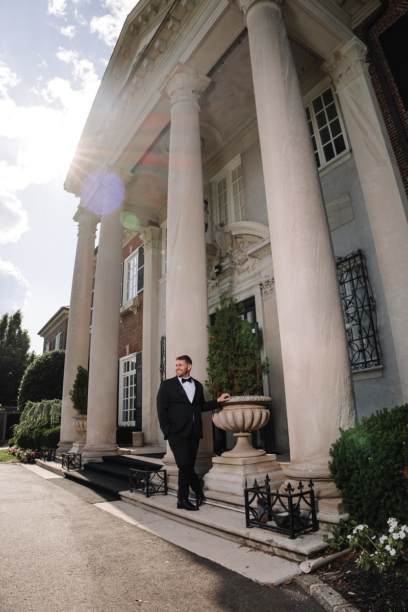 A groom in a black suit and kittel stands on a church building’s at The Mansion At Glen Cove, photographed by CJ Studios