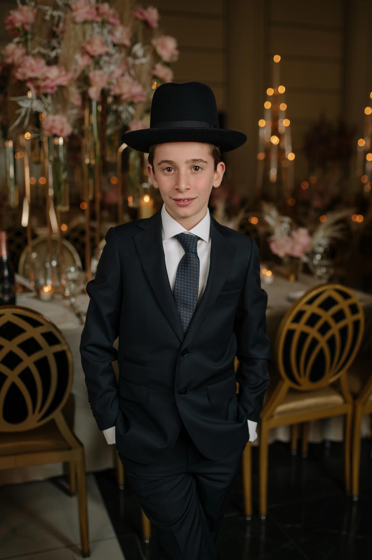 Framed portrait of a young Jewish boy in a black hat and suit with a tie, standing indoors at a decorated event with soft warm lights and floral centerpieces behind him.