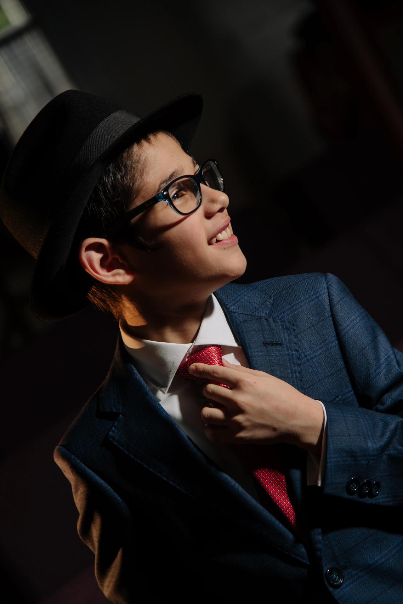 Young man in a blue suit and red tie wearing a black fedora and glasses, smiling as he looks upward during his celebration.