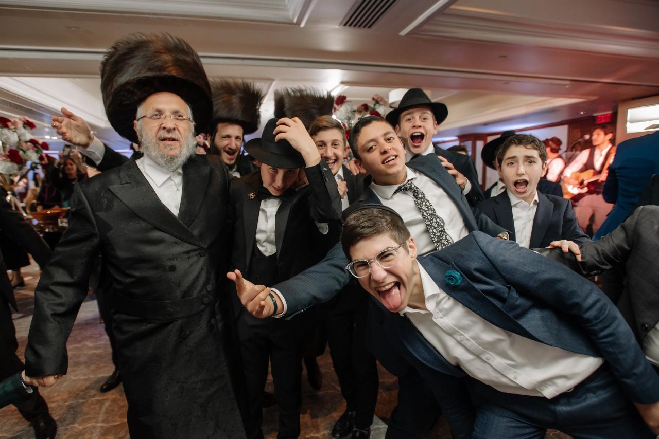 Smiling and cheering men in black hats and suits celebrate during a wedding dance, with one man in a dark suit pointing while others shout and laugh in the background.