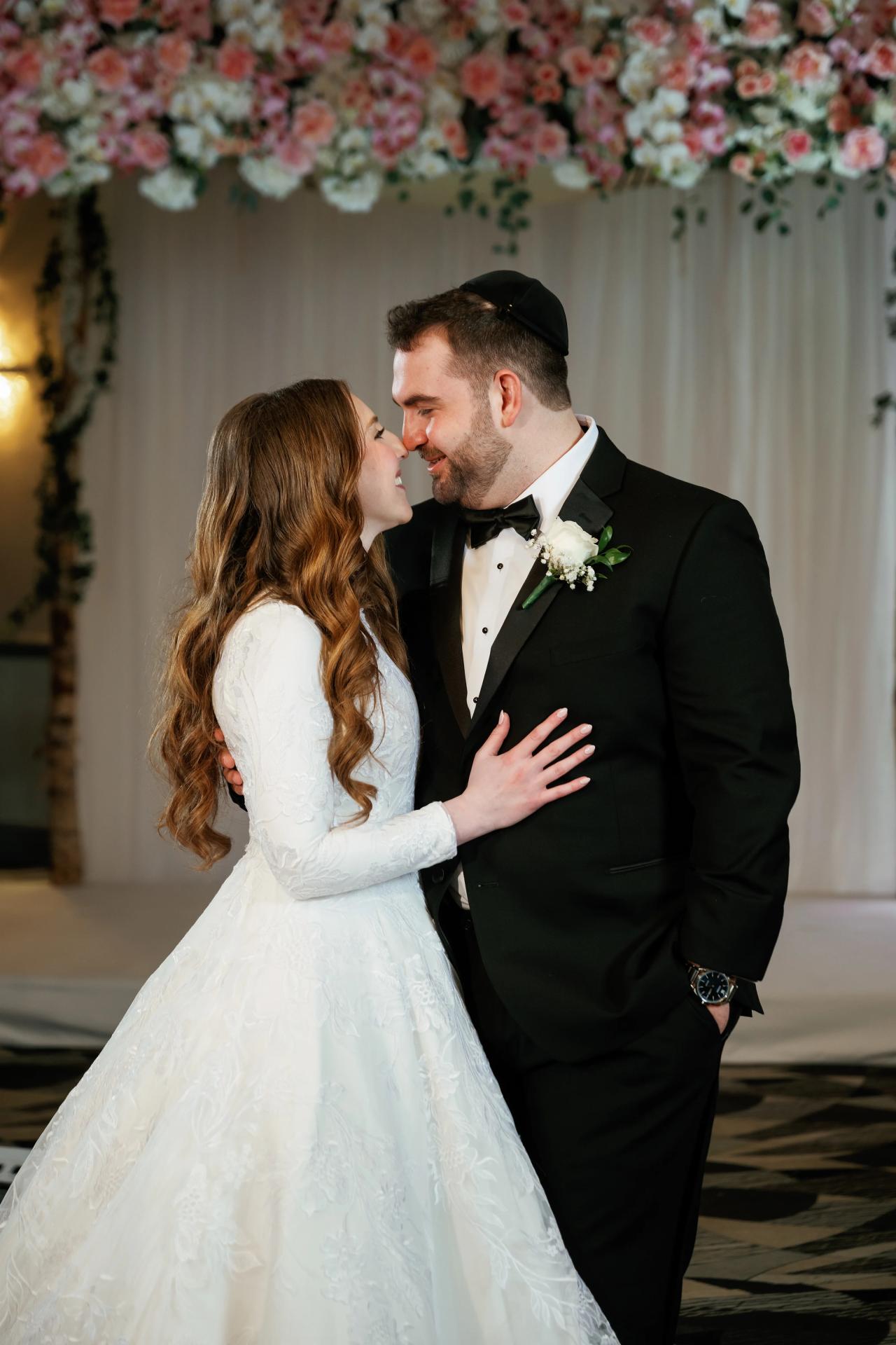 Bride and groom embracing in a close kiss moment during a ceremony under floral arrangements and white draping backdrop, warm lighting and soft bokeh.