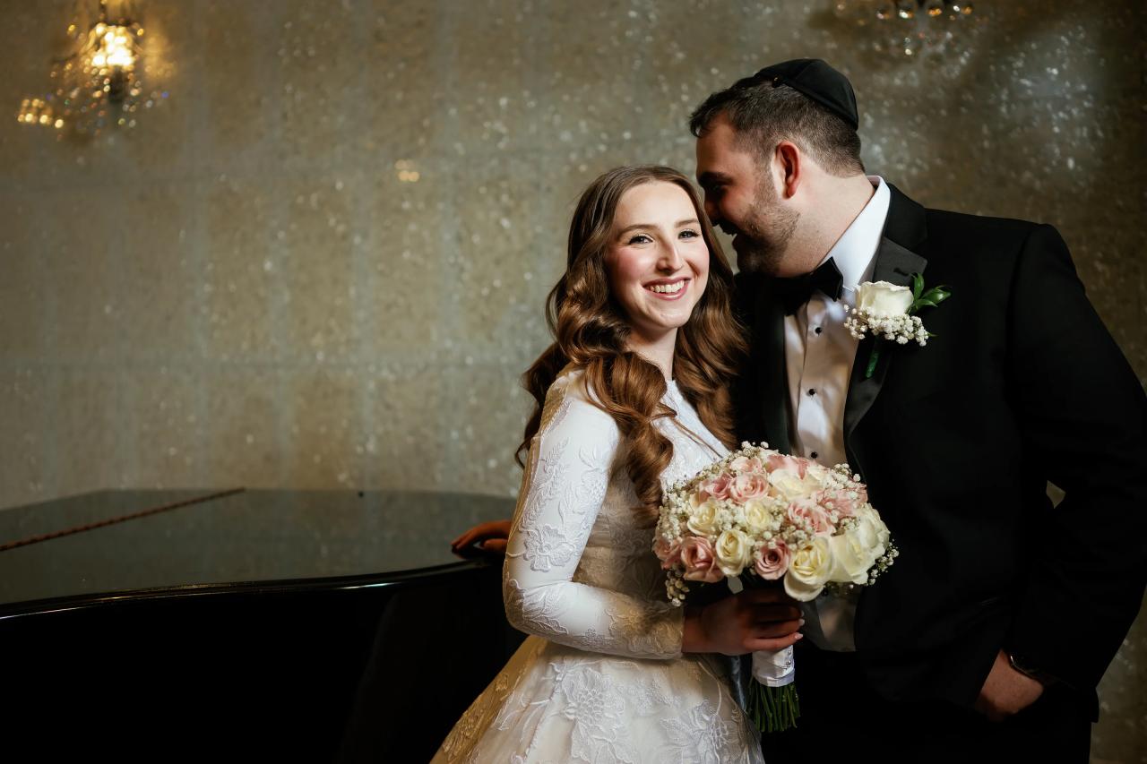 Warmly lit wedding portrait of a smiling bride and groom in a close embrace holding a bridal bouquet against a softly sparkling background.