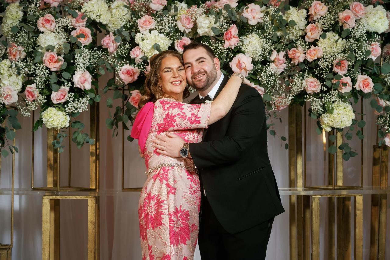 Bride and groom in a warm embrace, smiling for the camera in front of a richly floral backdrop with pink and cream flowers.