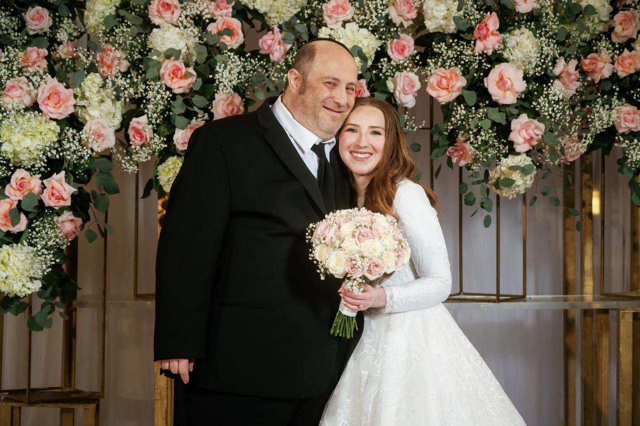 Bride smiling beside groom in an intimate embrace, holding a soft pastel bouquet, in front of a floral chuppah backdrop with roses and greenery.