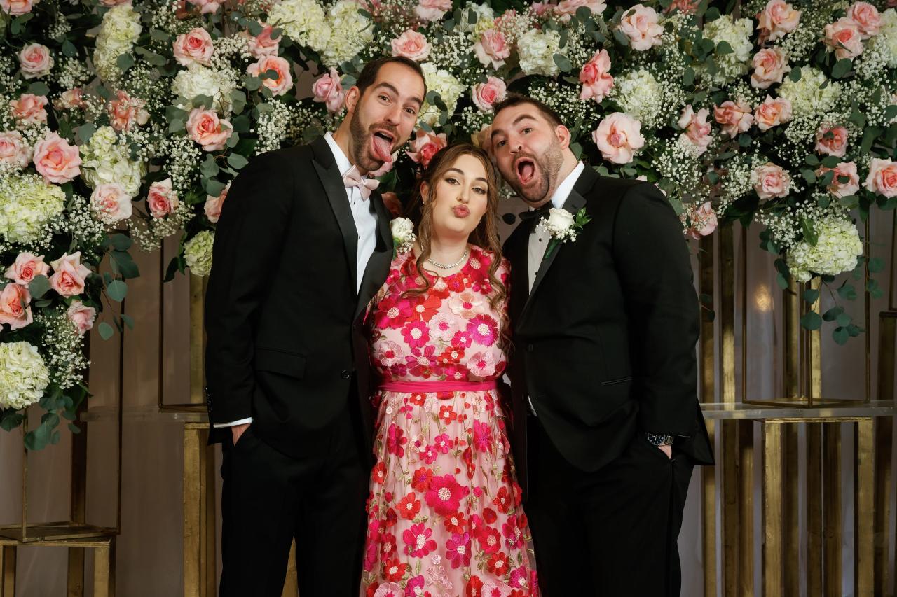 Bride centered between two men in black suits, smiling with playful open-mouth expressions in front of a pink-and-white floral backdrop.