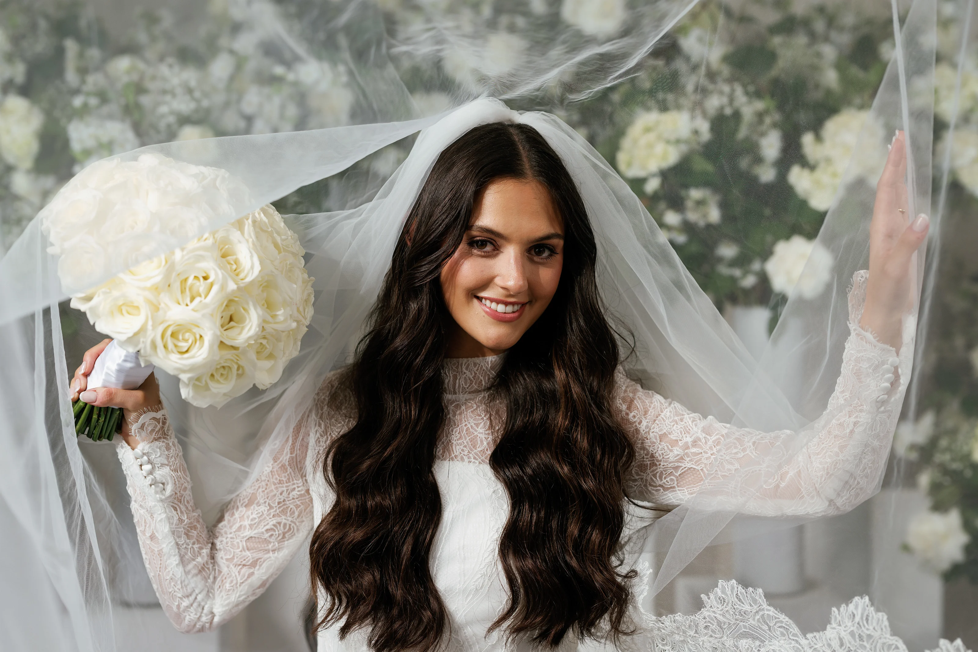 Portrait of a bride smiling under a flowing white veil while holding a bouquet of white roses against a floral backdrop.