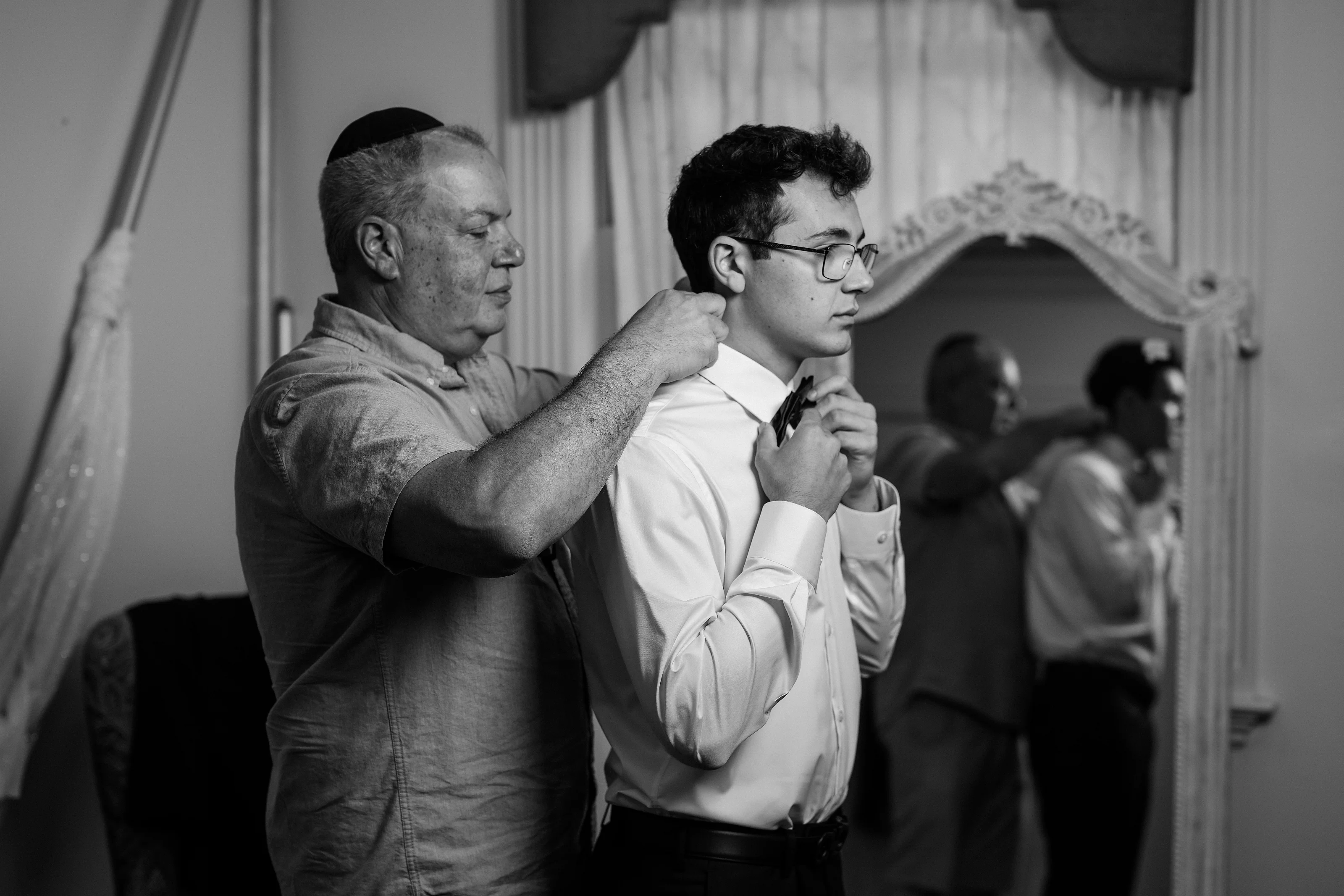 Black-and-white candid photo of an older man helping adjust the groom’s garment during a tefillin moment, with guests blurred in the background.