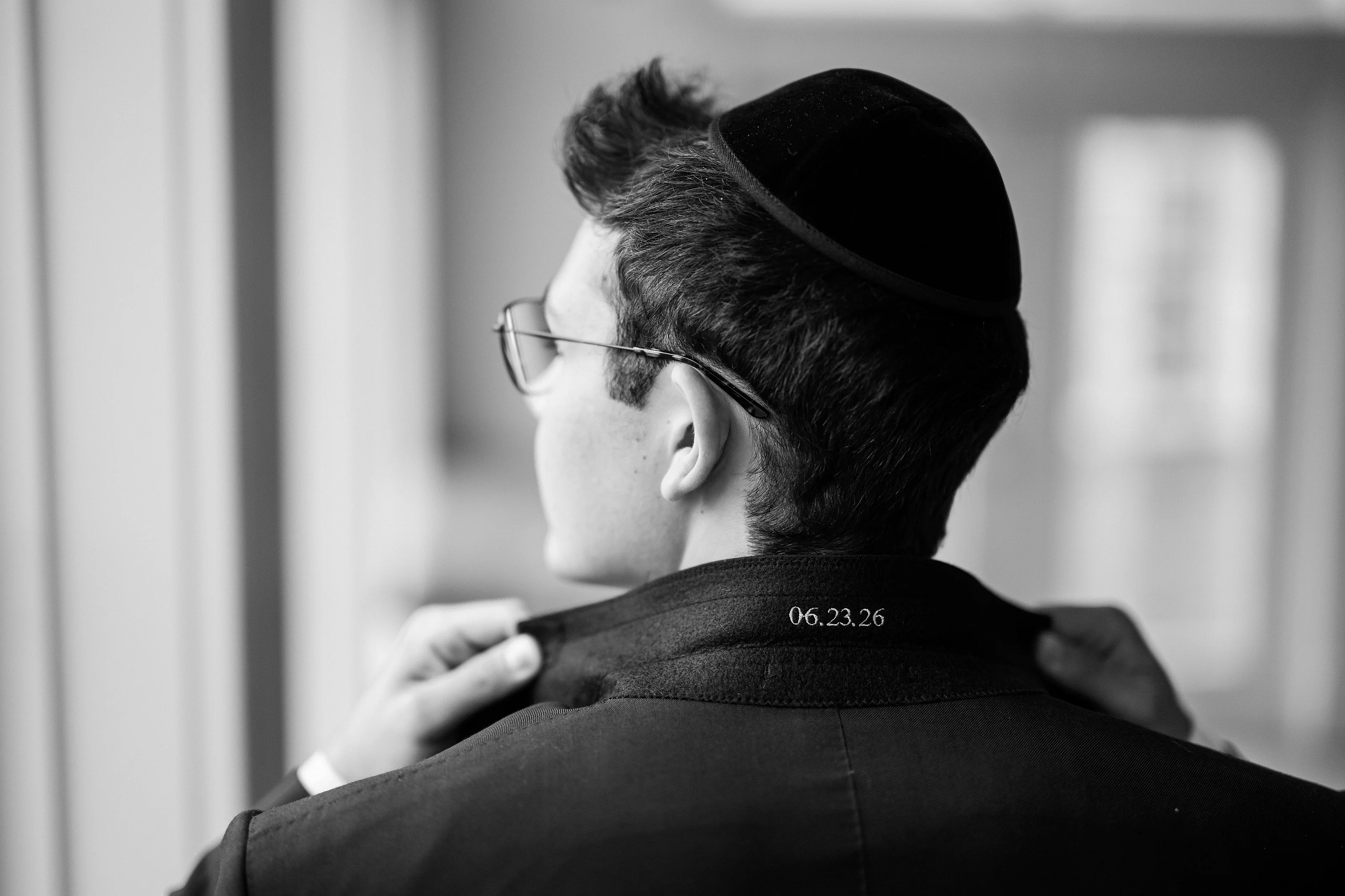 Black-and-white profile portrait of a man in a black kippah and glasses during kabbalas-panim, hand resting on his shoulder.