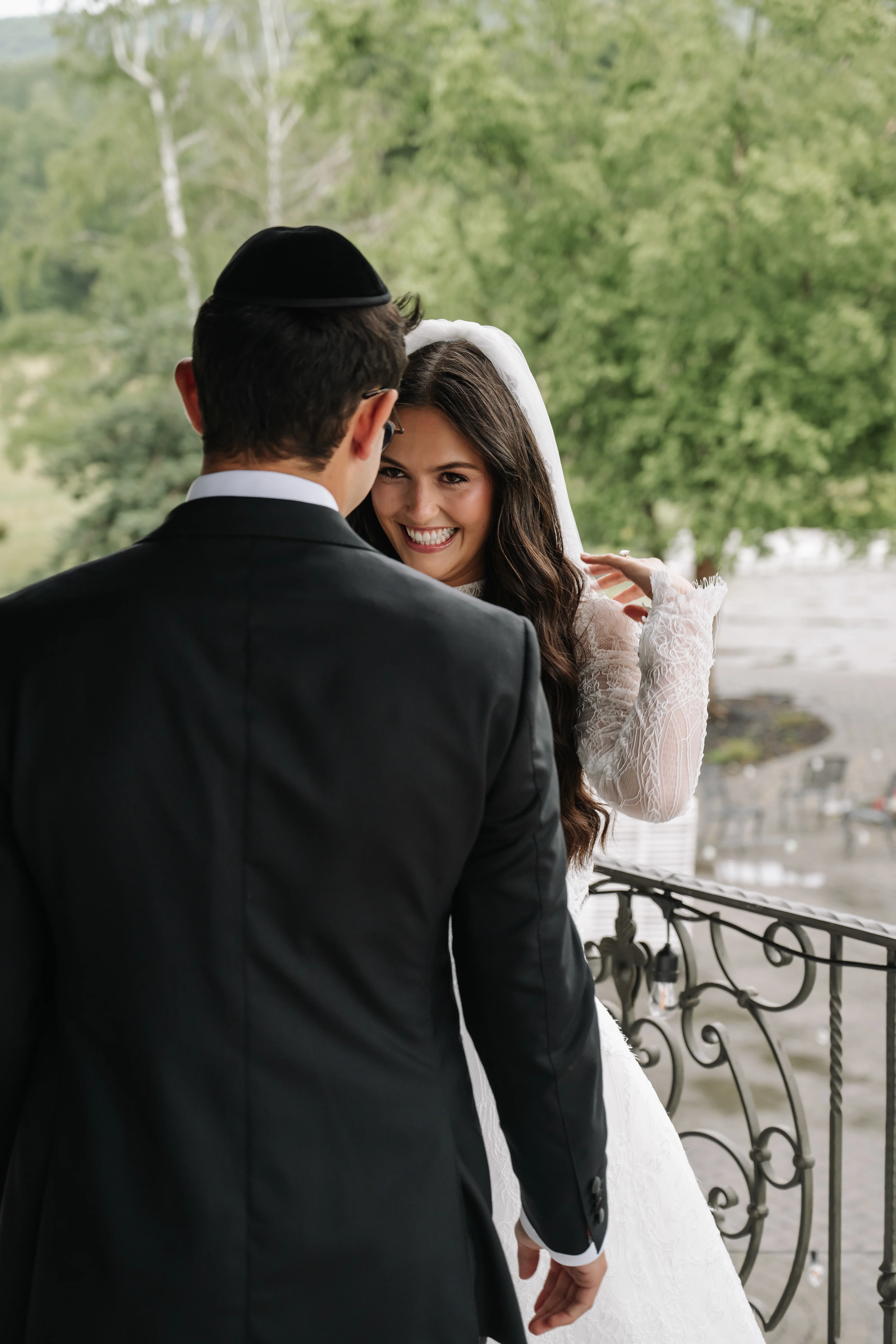 Bride smiling under a white veil as she leans toward her groom in a black suit during a wedding badeken moment on a balcony with greenery in the background.