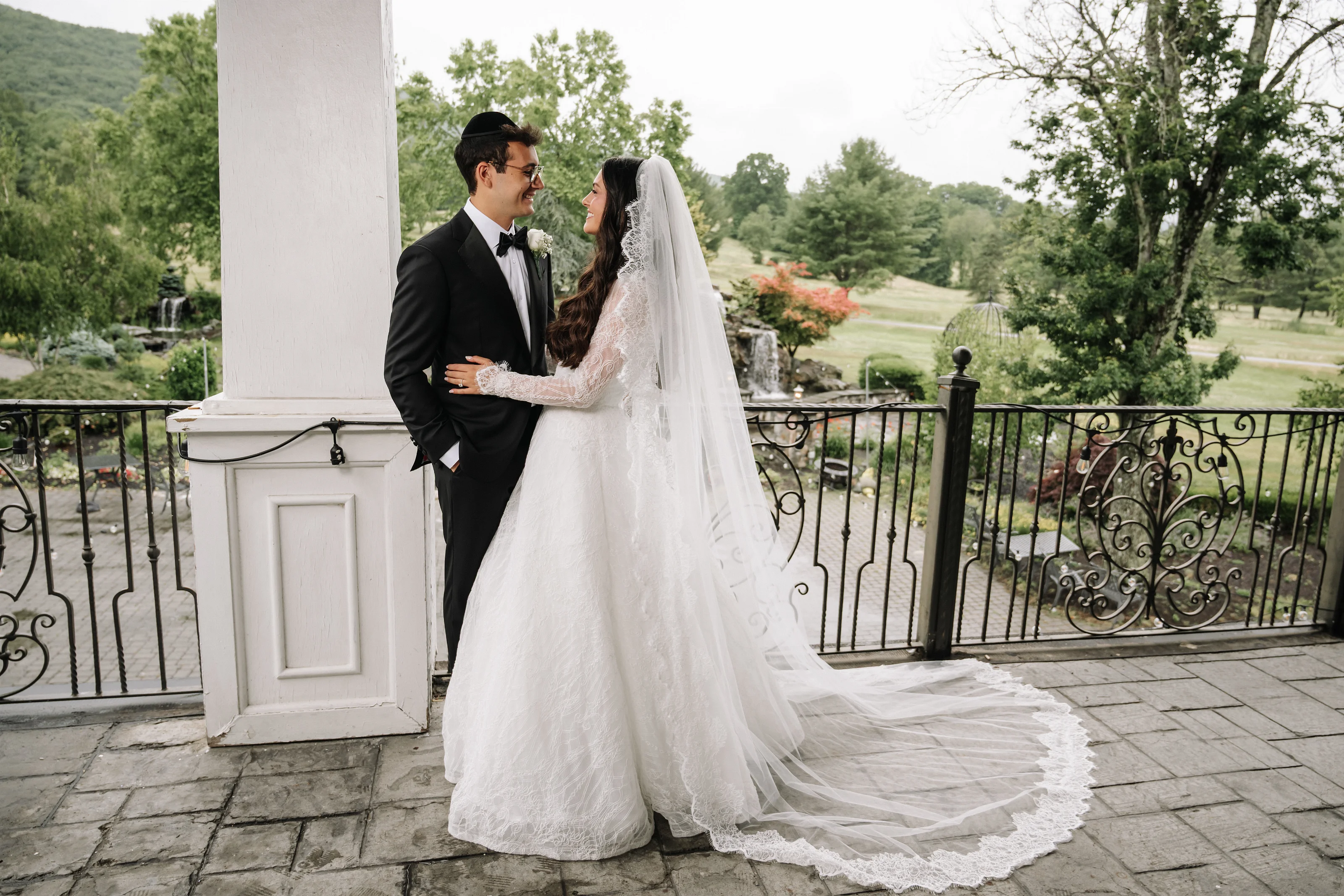 Bride and groom embracing on a terrace, gazing at each other with soft smiles under a veil in an outdoor garden setting.