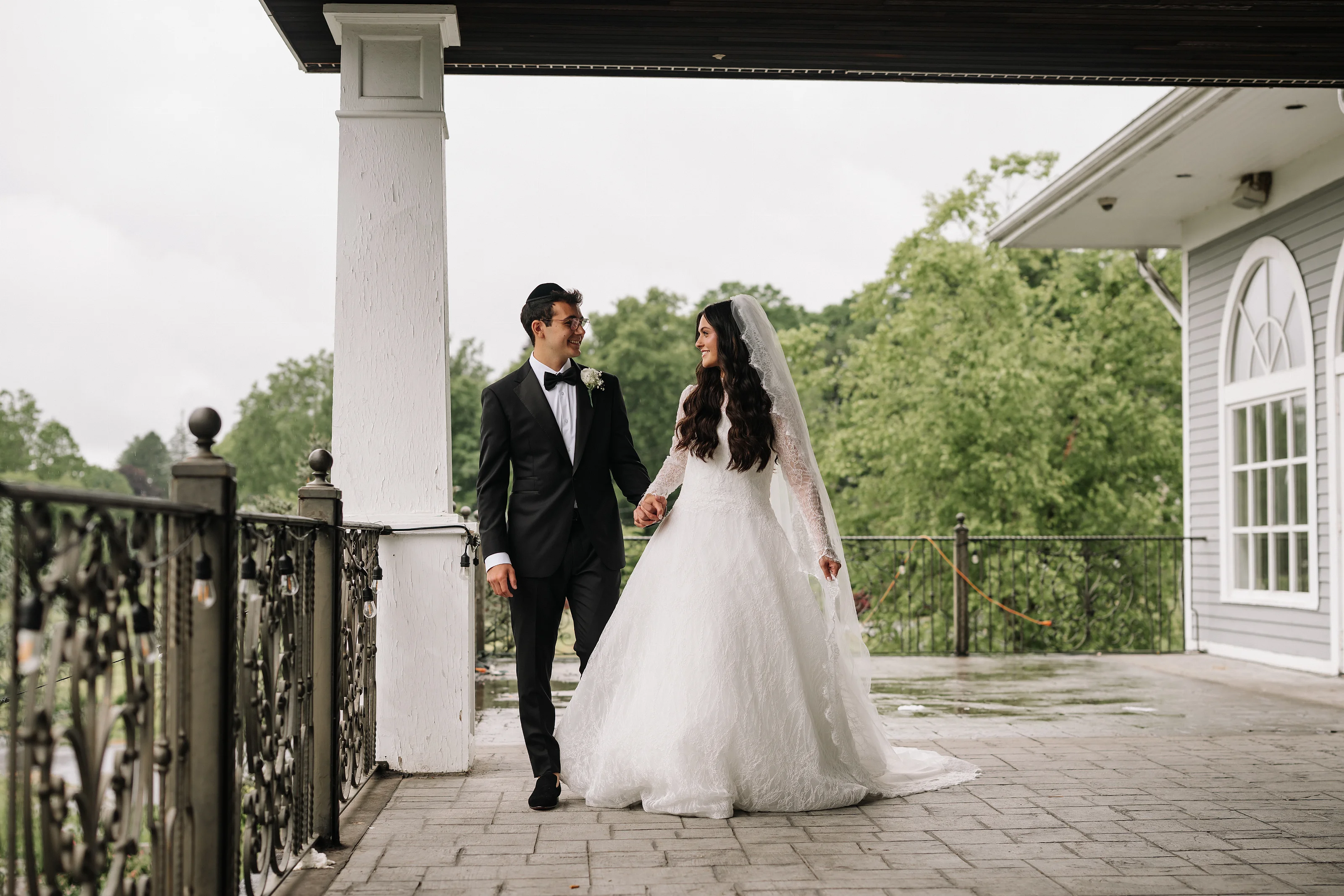 Wedding couple holding hands and walking together outdoors on a porch walkway, bride in a lace veil and gown beside the groom in a black tuxedo.