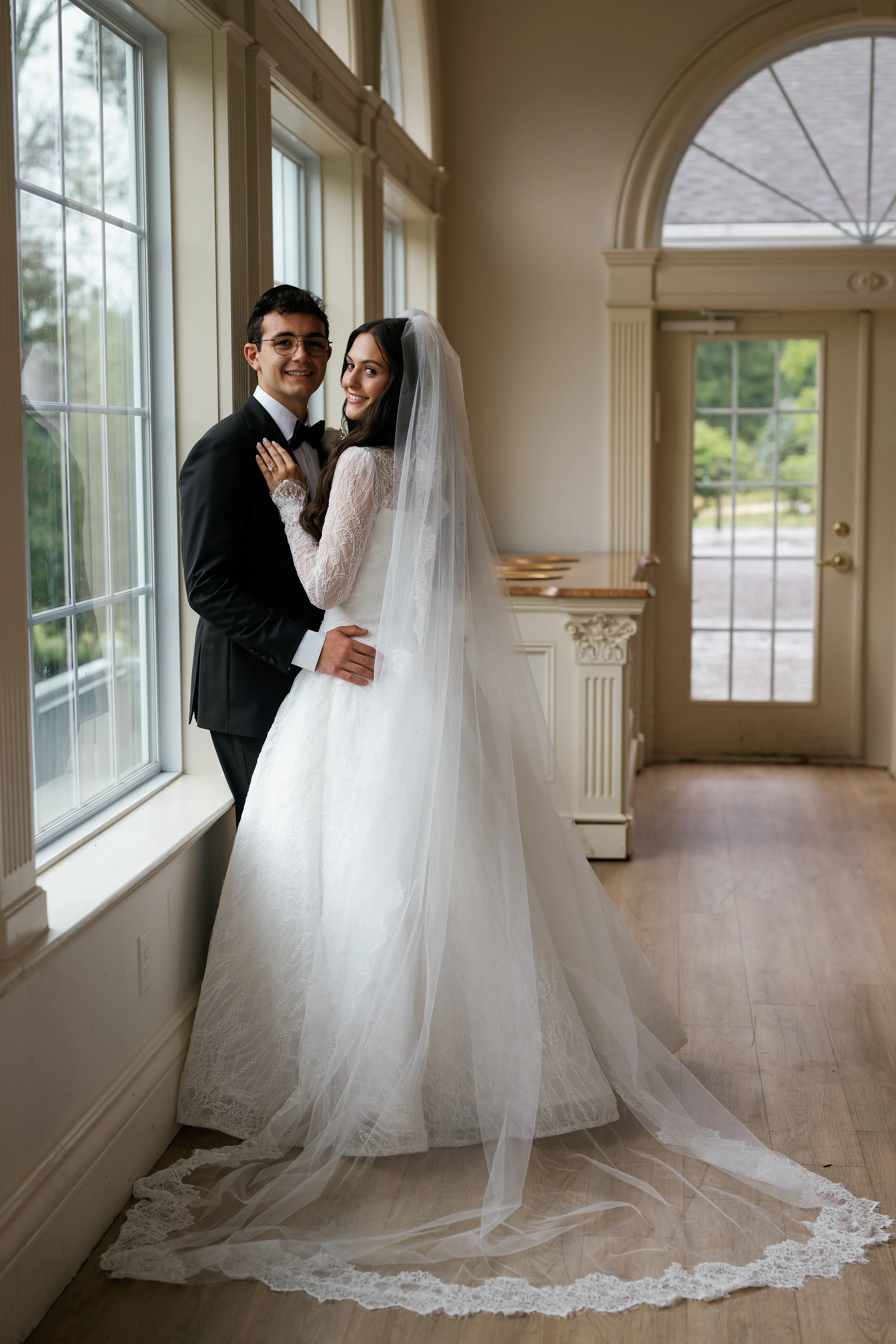 Bride and groom in an embrace by tall windows, bride in a lace dress and veil with warm natural light inside a bright arched room.