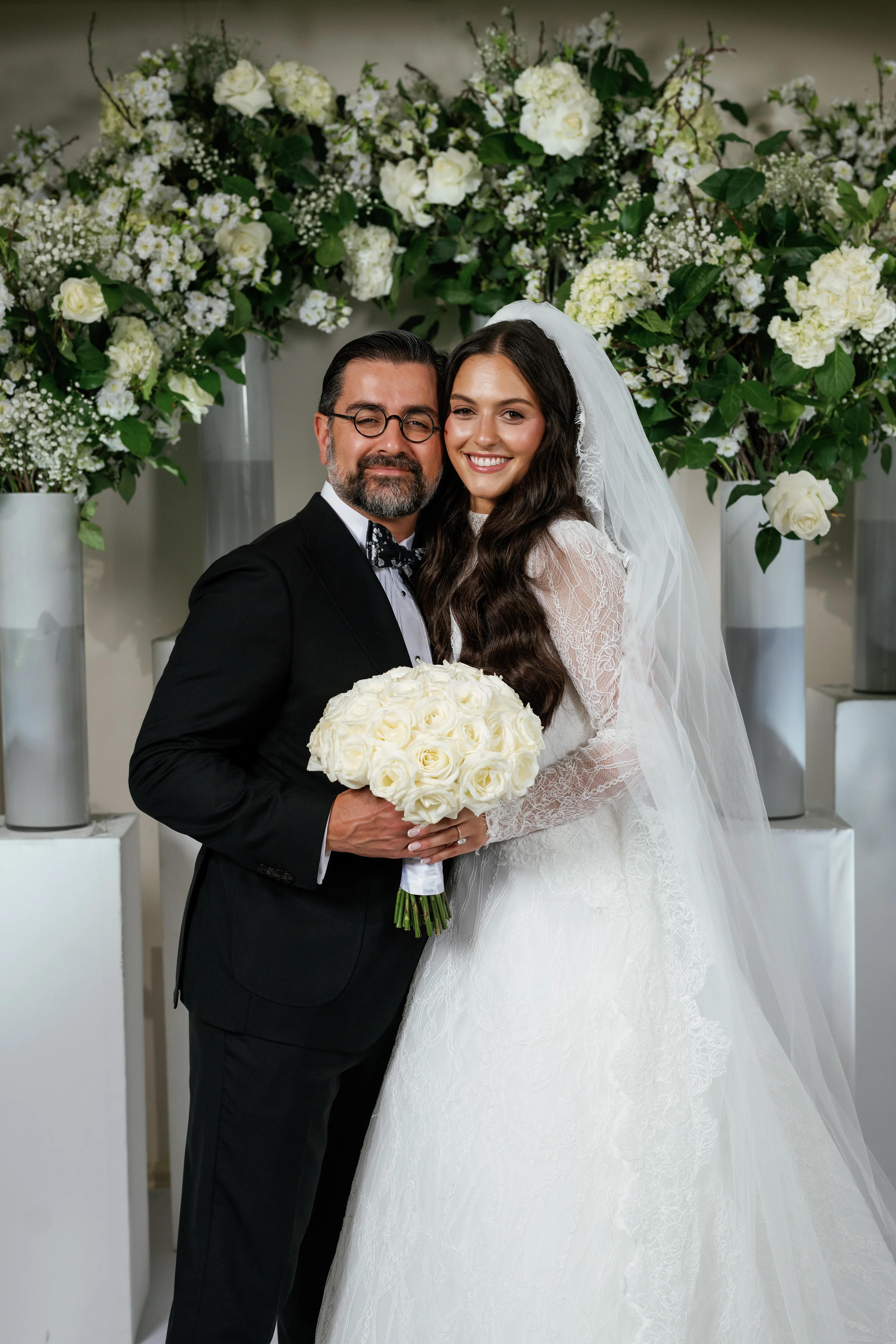 Smiling bride and groom embracing and holding a white rose bouquet in front of a wall of white flowers and greenery.