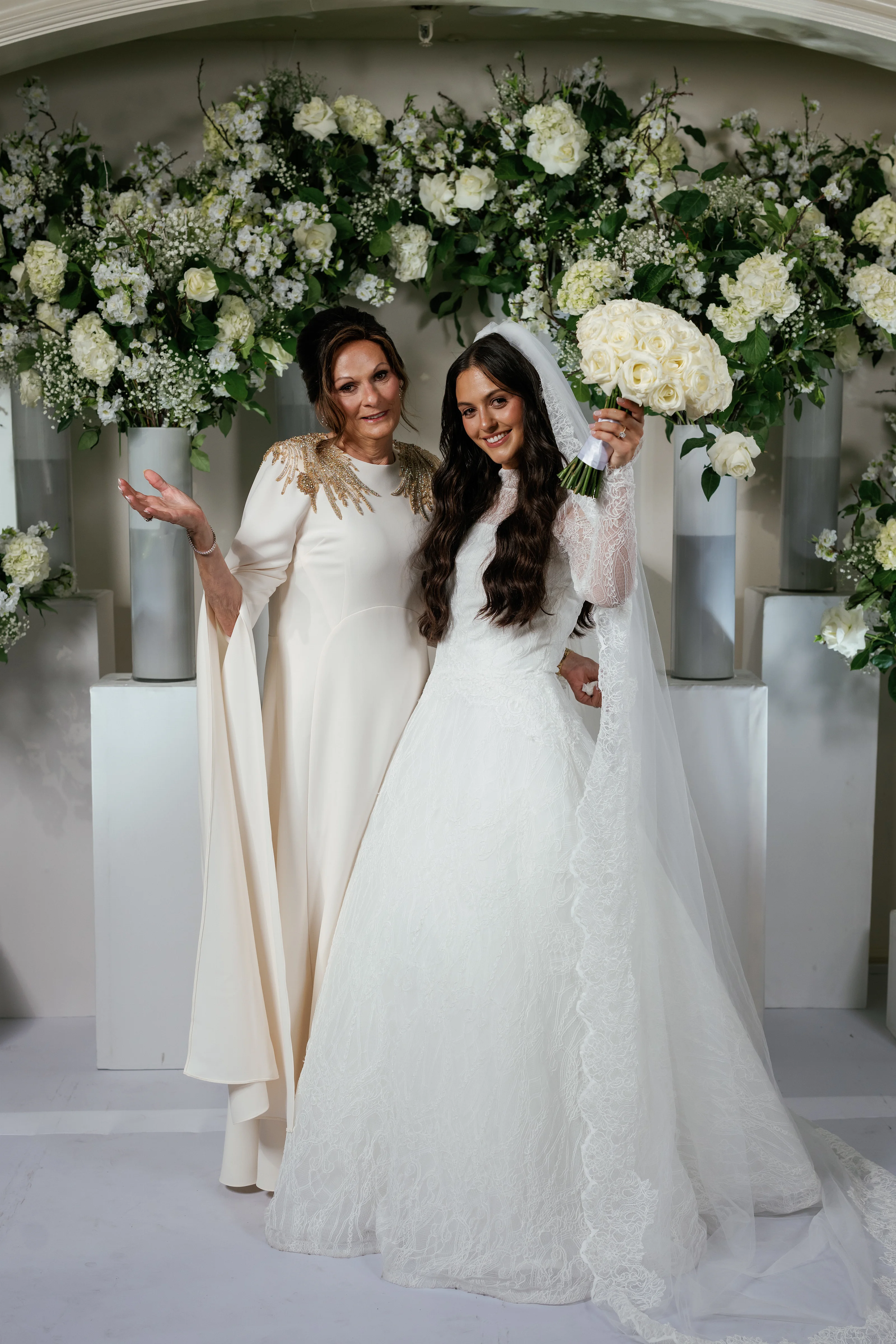 Bride in a white gown and veil smiling beside a loved one under a floral chuppah, holding a bouquet as they pose for a wedding portrait.