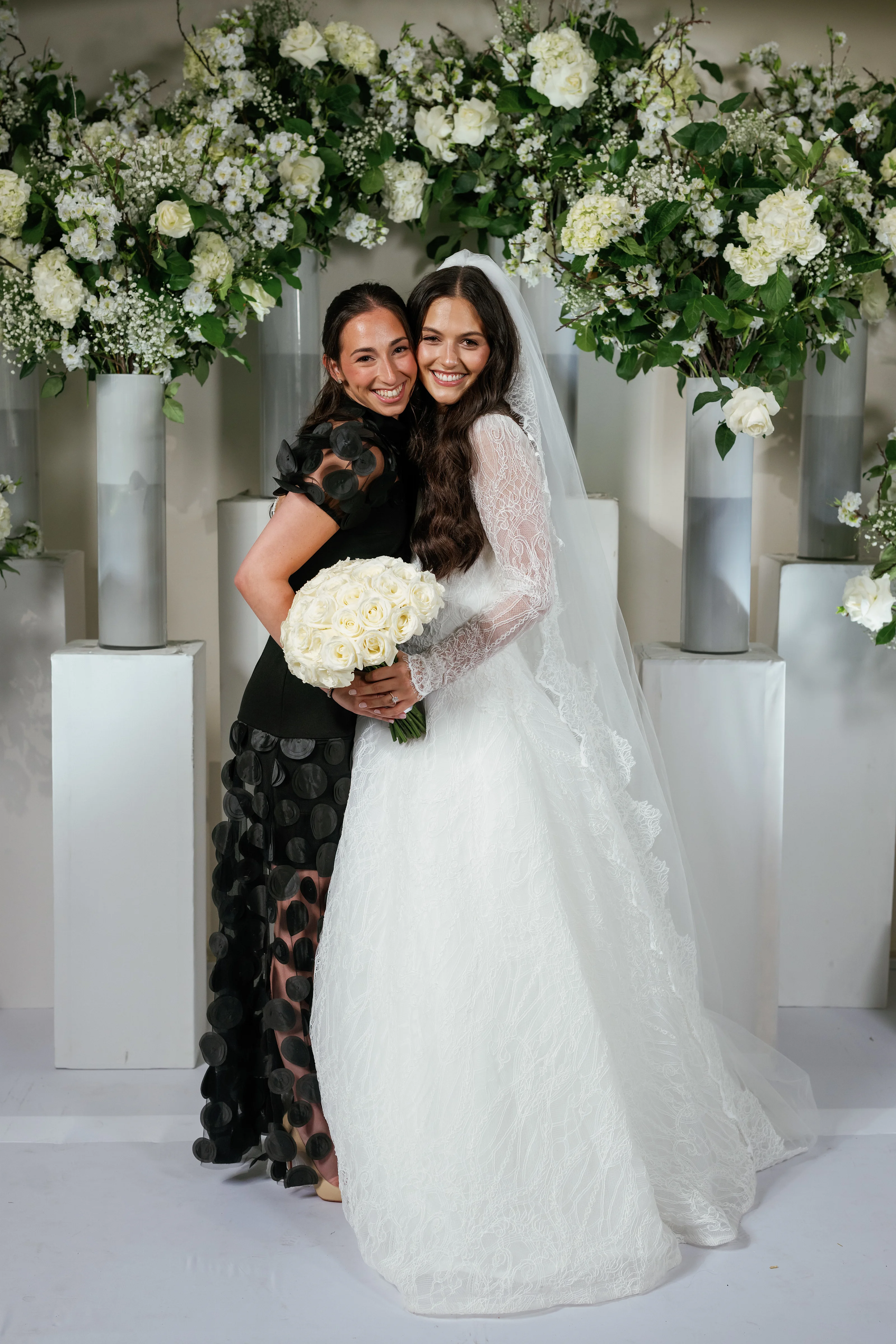 Smiling bride in a white gown and veil embracing a close companion in front of white floral arches and pedestal displays, holding a white bouquet.