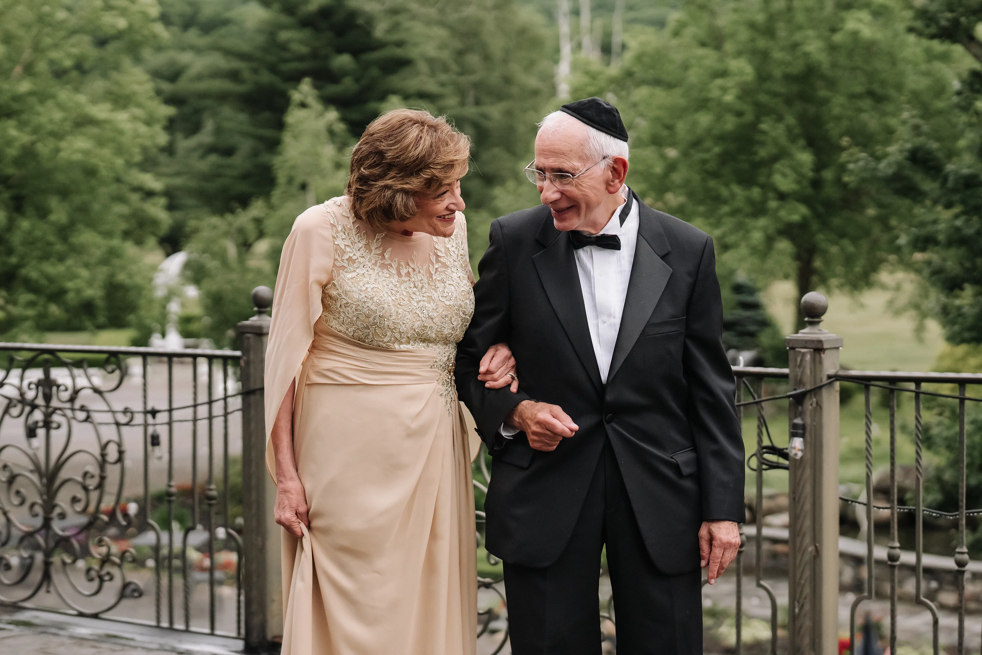 Bride and groom smiling and holding hands during an emotional embrace outdoors near an iron fence with trees in the background.