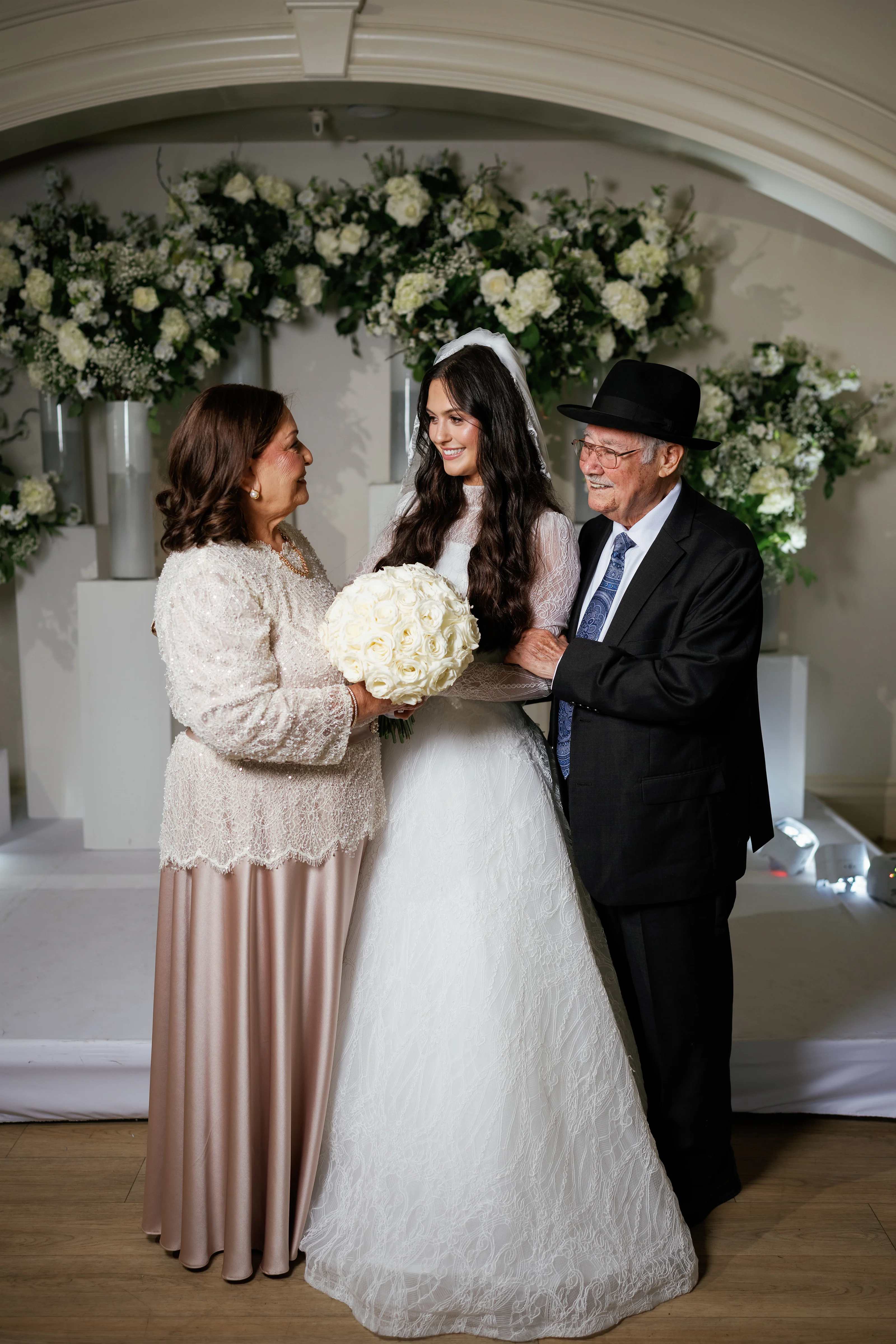 Bride holding a white floral bouquet during badeken, standing between family in front of an arched floral chuppah backdrop.