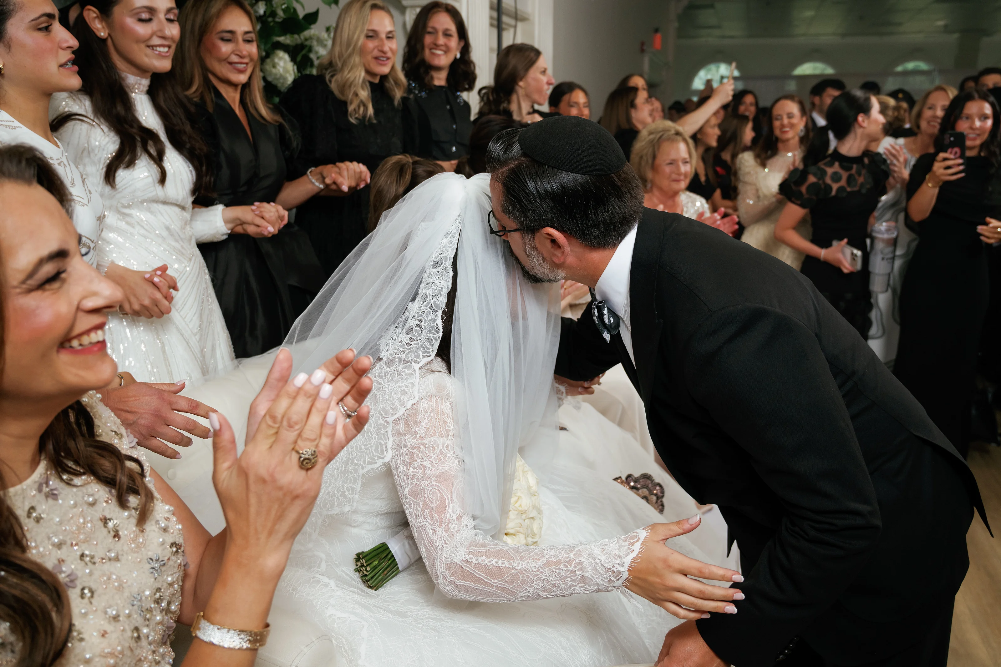 Bride and groom during kabbalas-panim as guests smile, clap, and reach toward them in an event hall.