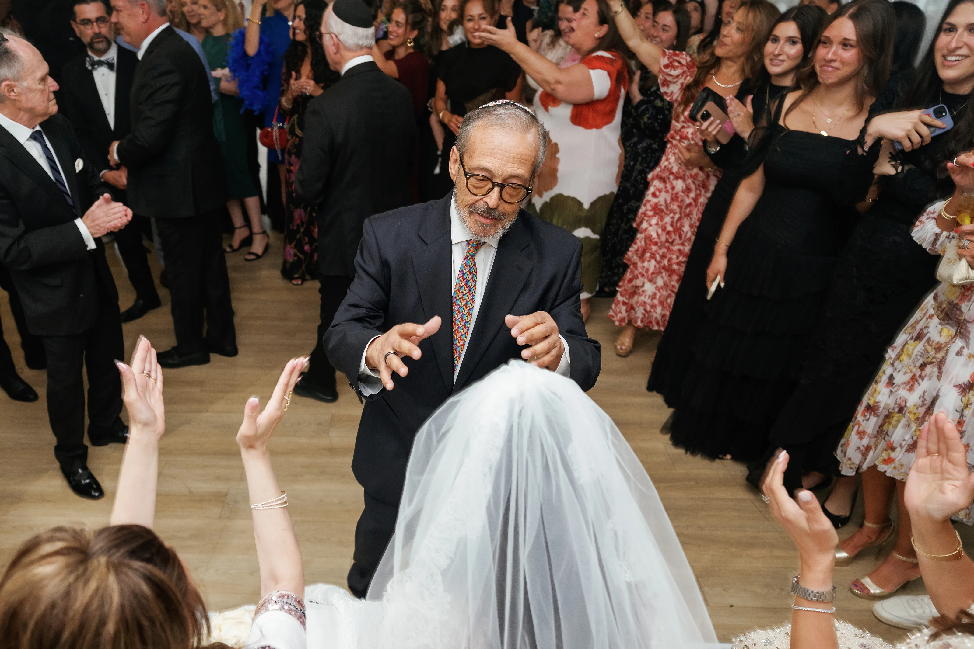 A man in a suit dances with expressive hands while guests clap and cheer around him at an indoor wedding celebration.