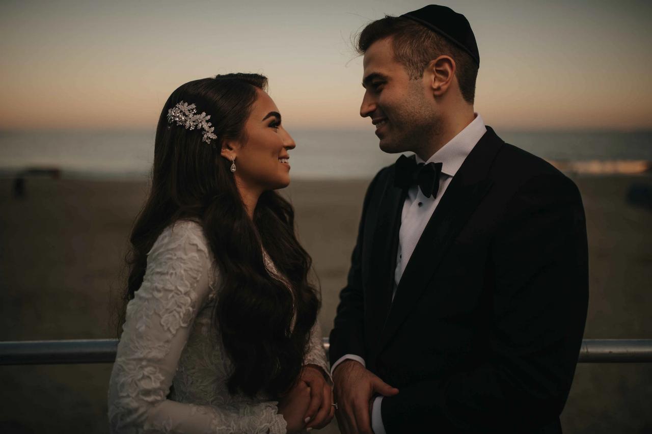 Frum wedding couple in formal attire holding hands and smiling at each other during sunset, bride in a jeweled headpiece and groom in a black tuxedo with bow tie.