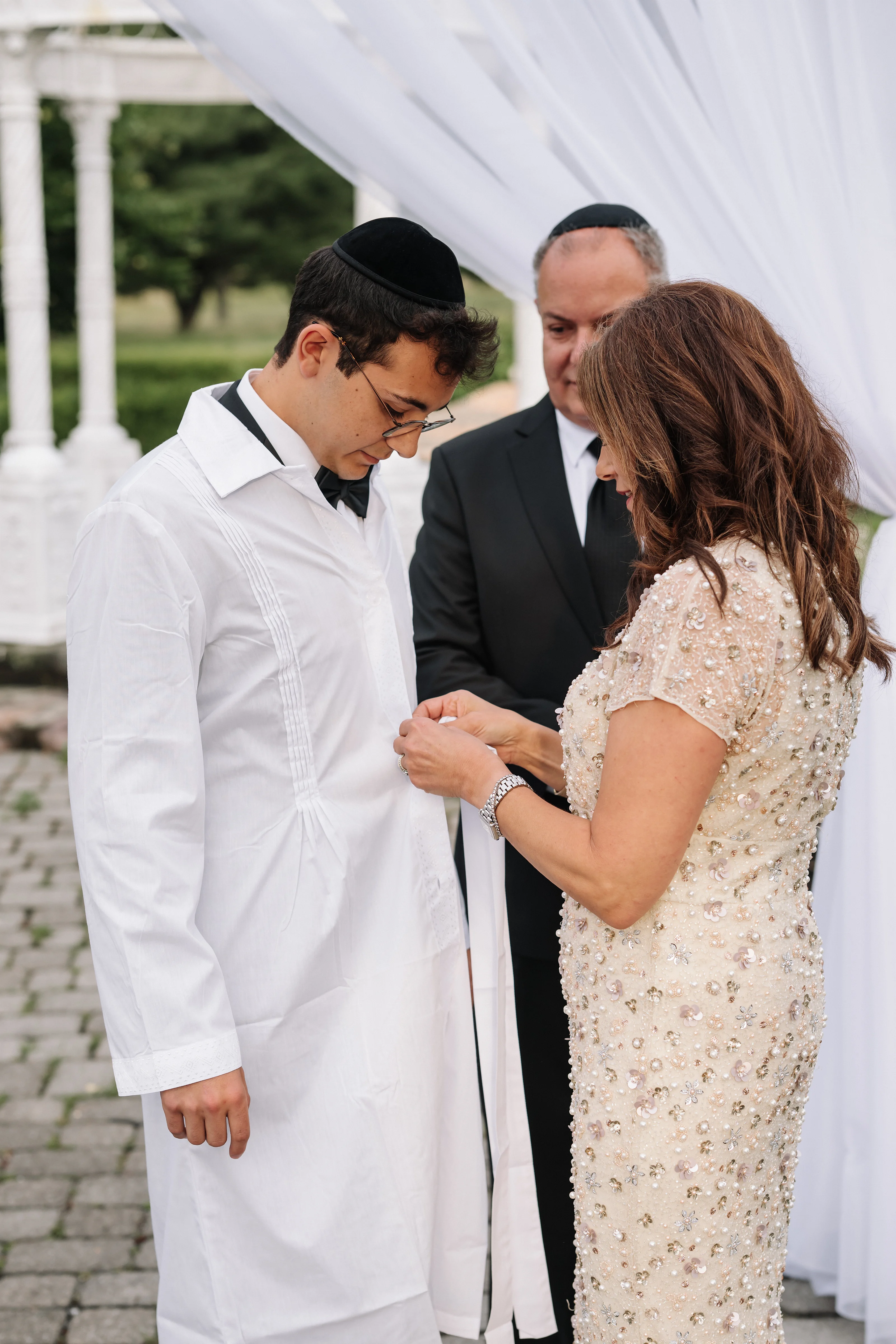 Bride in an embellished gown and groom in a black hat share an intimate close moment under a white canopy, with their hands gently meeting.