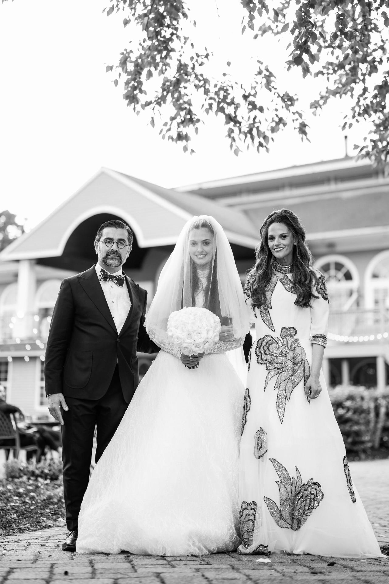 Black-and-white wedding portrait of the bride walking forward in her veil and gown with the groom and a family member outdoors.
