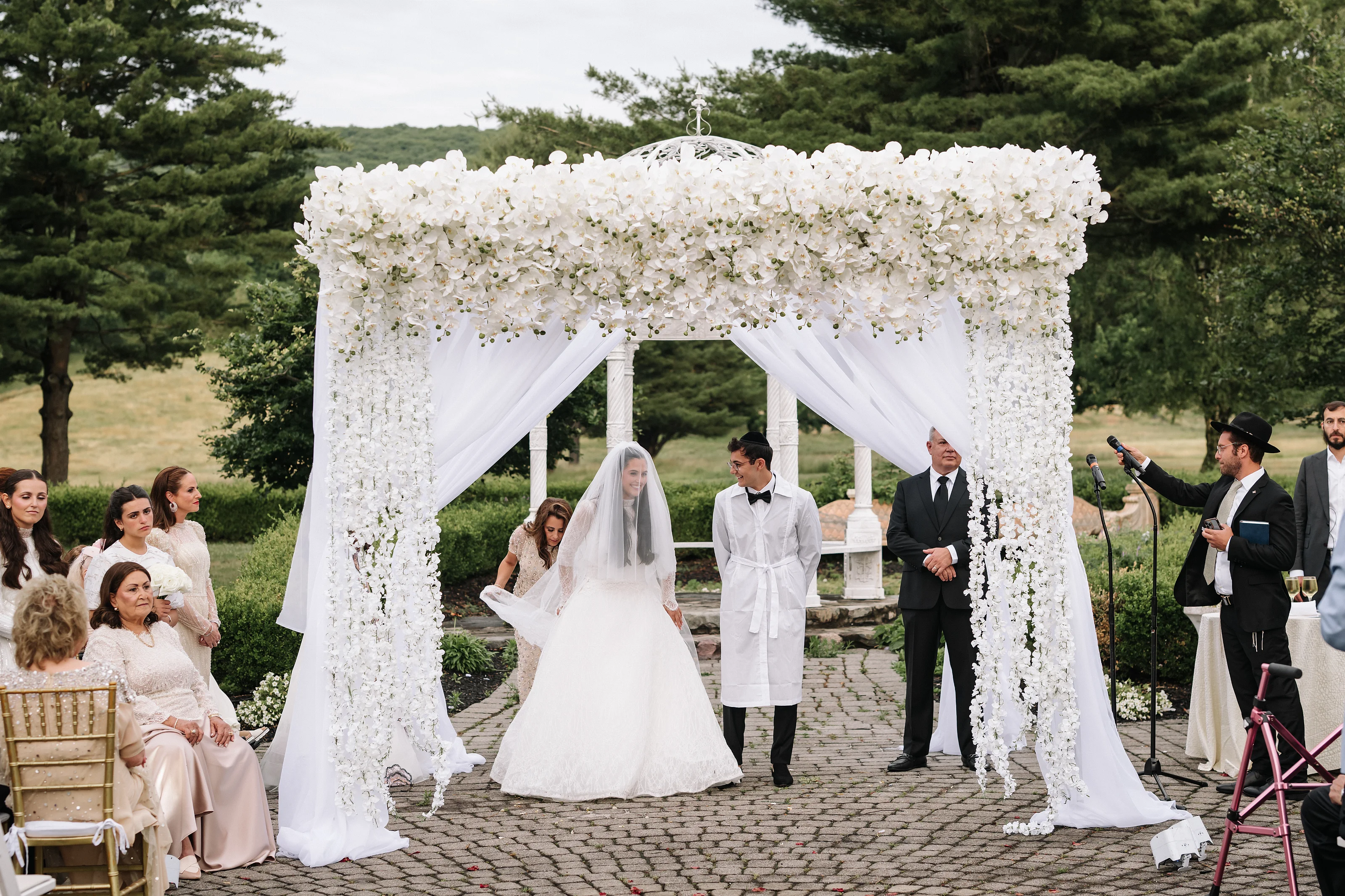 Wedding ceremony under a white floral chuppah outdoors, with the bride walking forward as the groom and officiant stand beneath the canopy.