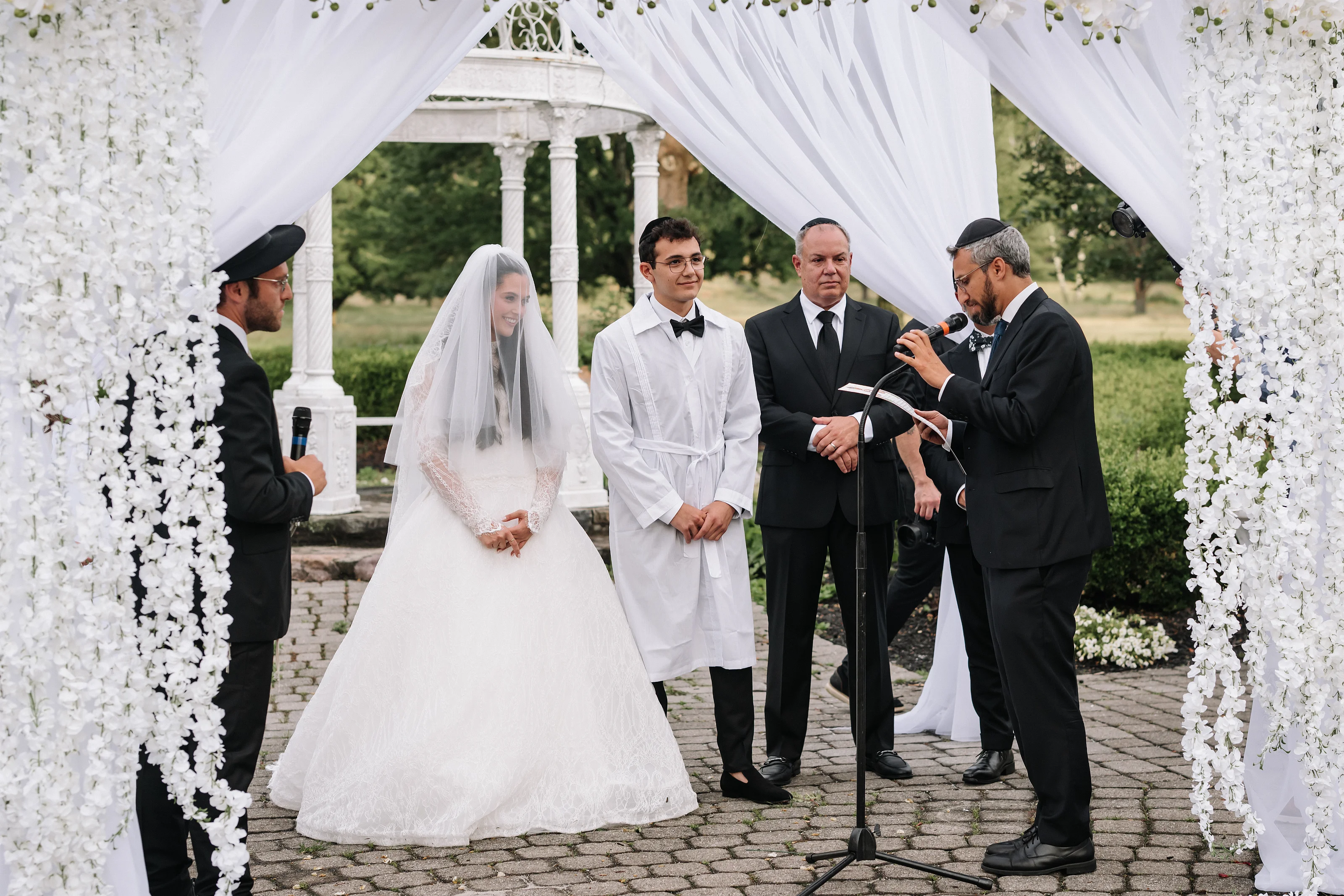 Bride and groom-side attendants under a white chuppah canopy during a ceremonial moment, with a speaker holding a microphone.