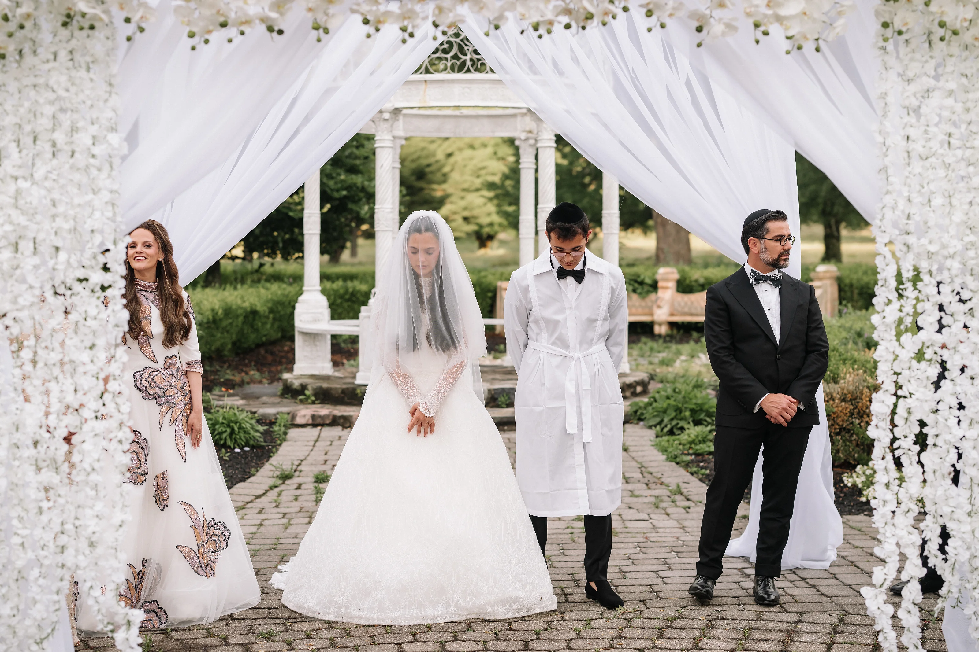 Bride in a white veil under a floral canopy with the groom beside her during an outdoor Jewish wedding ceremony.