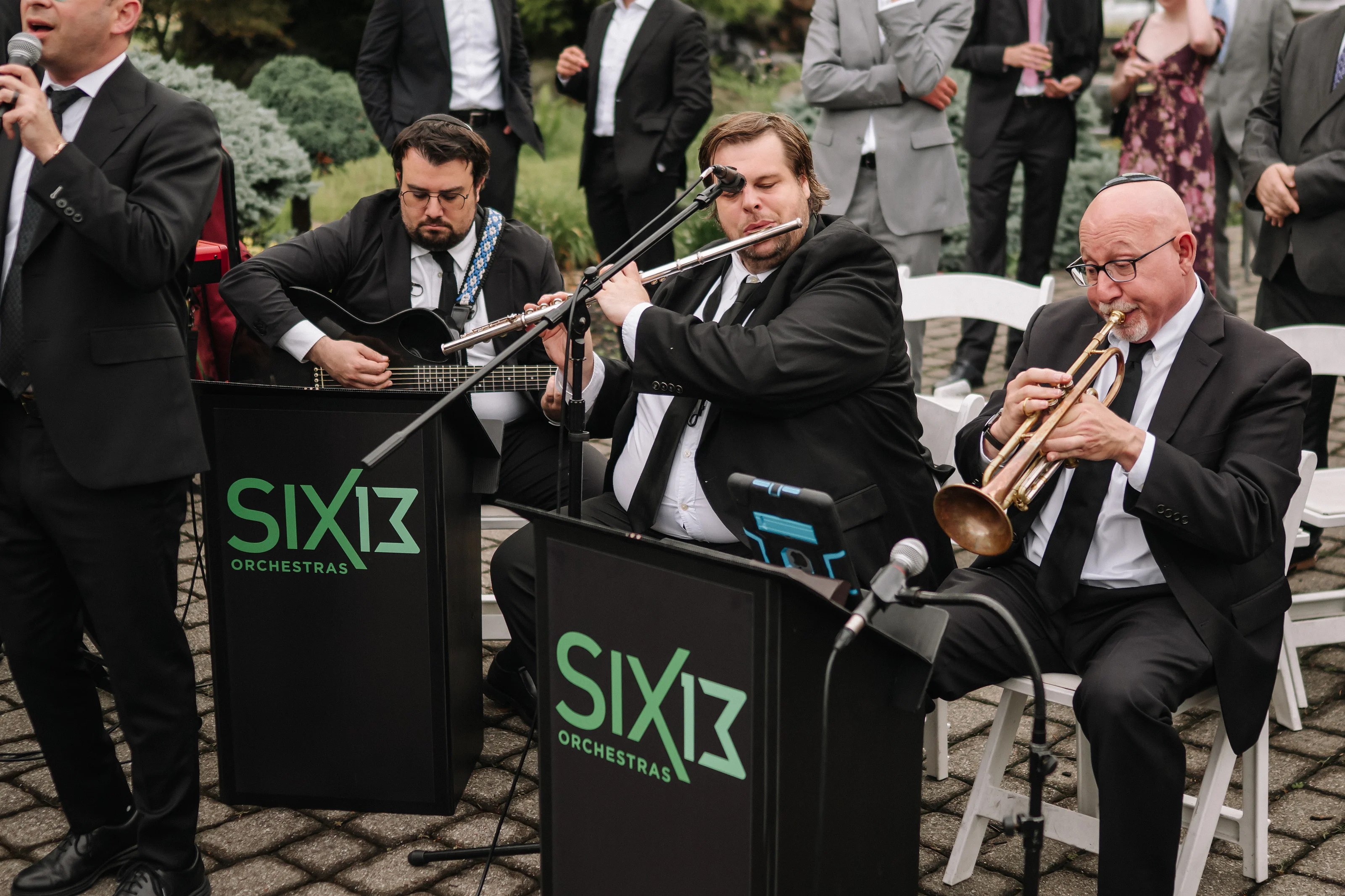 Outdoor wedding band performing with trumpeter and flutist in front, microphones and seated musicians, guests standing in the background.