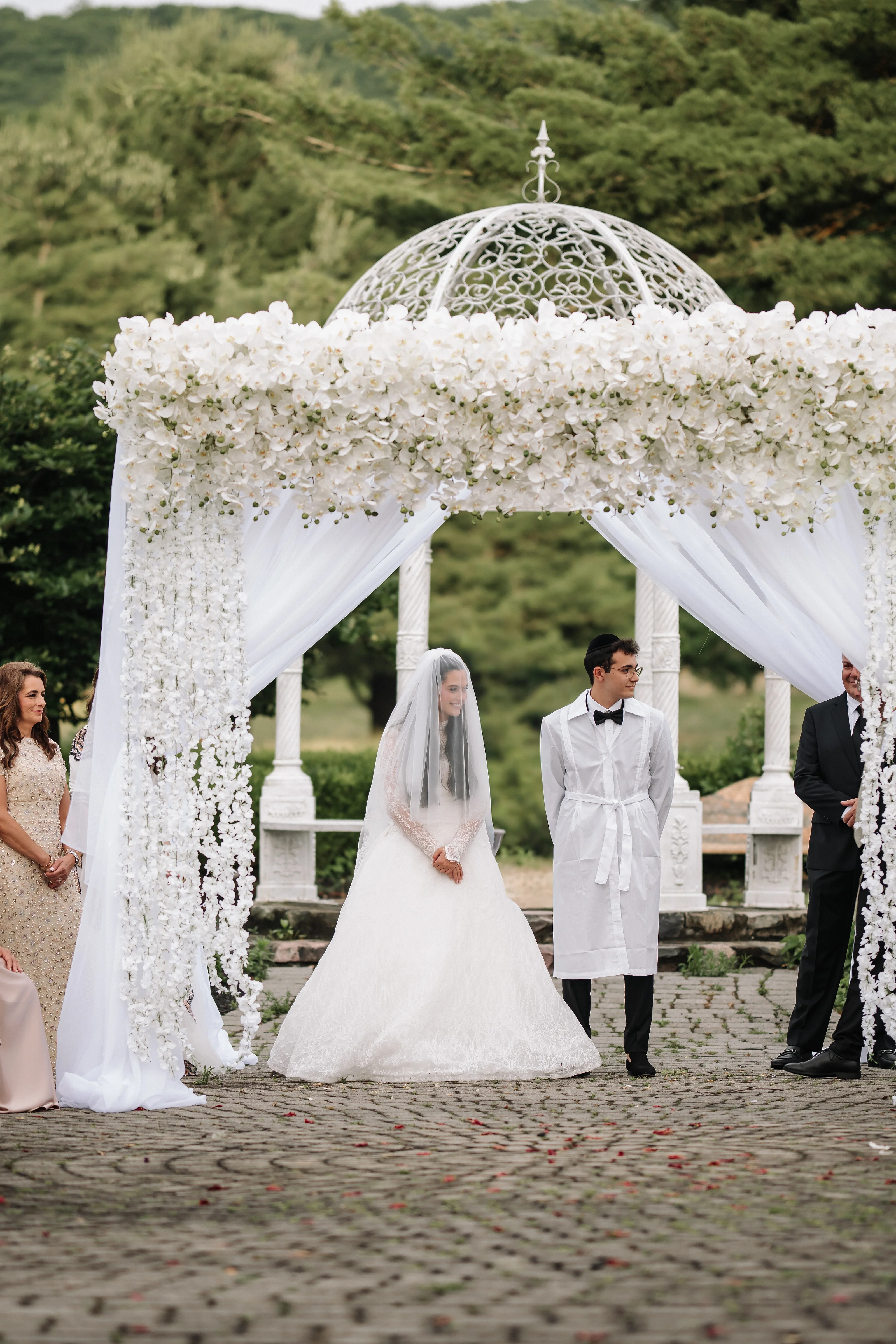Bride and groom walking under an ornate white floral wedding canopy during an outdoor ceremony.