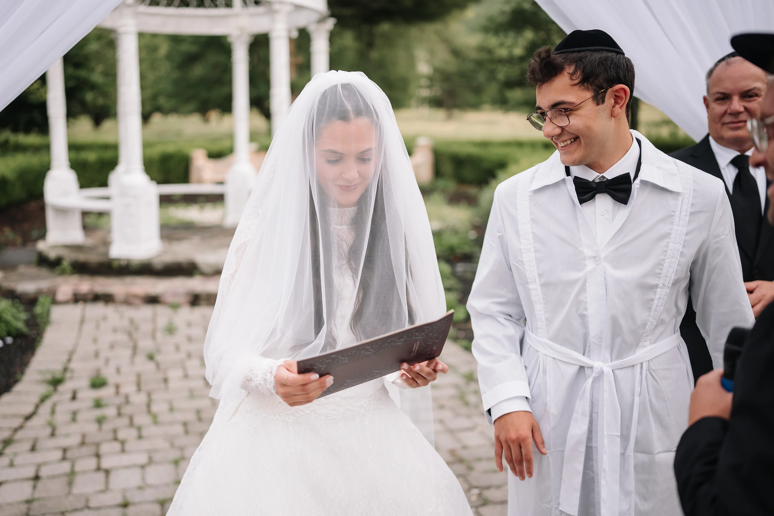 Veiled bride reading a ketubah while the groom in a black kippah smiles during badeken under a white canopy.