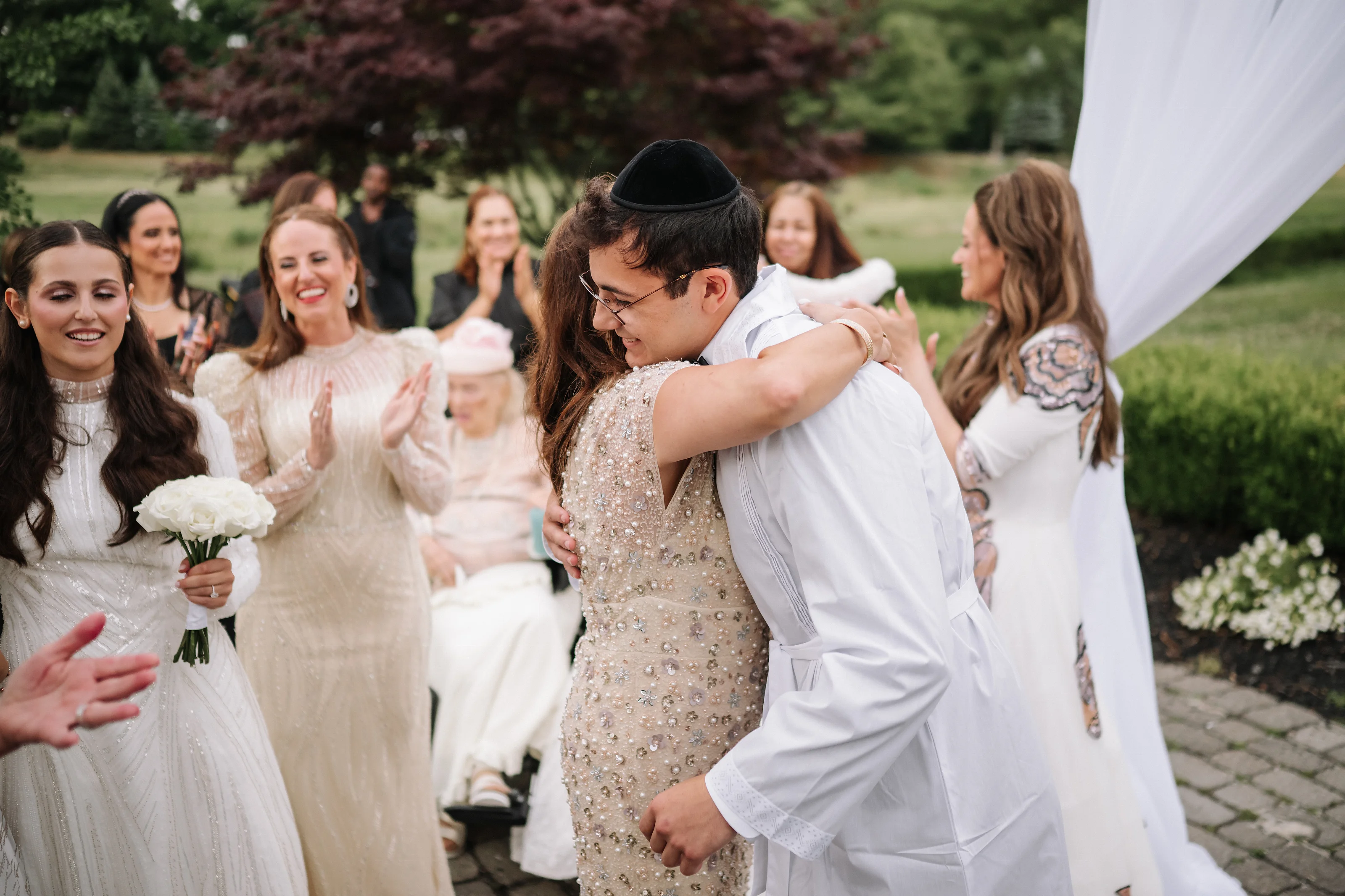 Bride and groom sharing a joyful hug outdoors while family members smile and clap in the background.