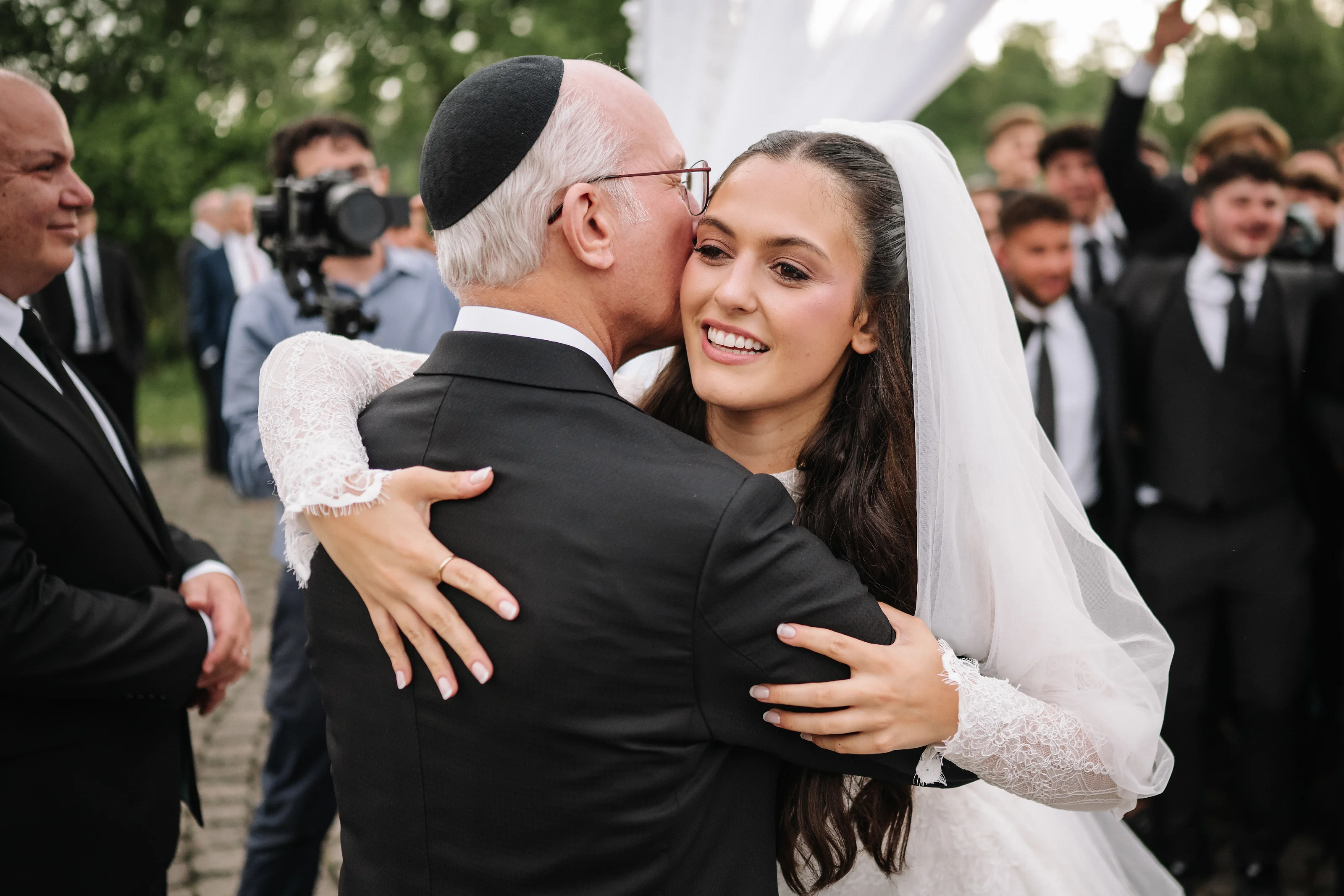 Bride in a white veil embracing her groom outdoors, joyful smile during badeken while guests and photographers blur in the background.