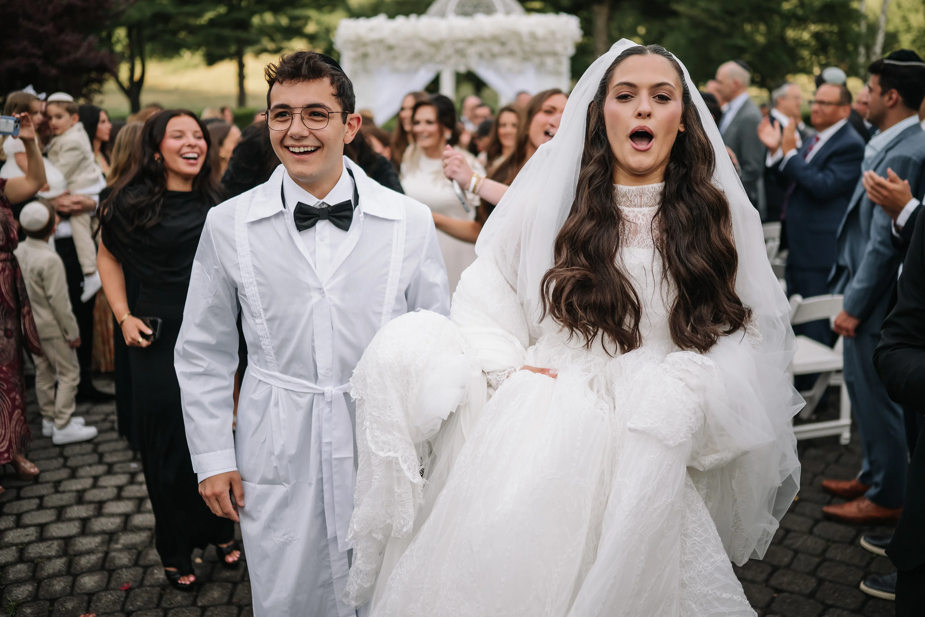 Bride and groom smiling and laughing outdoors during the celebration as guests watch in the background.