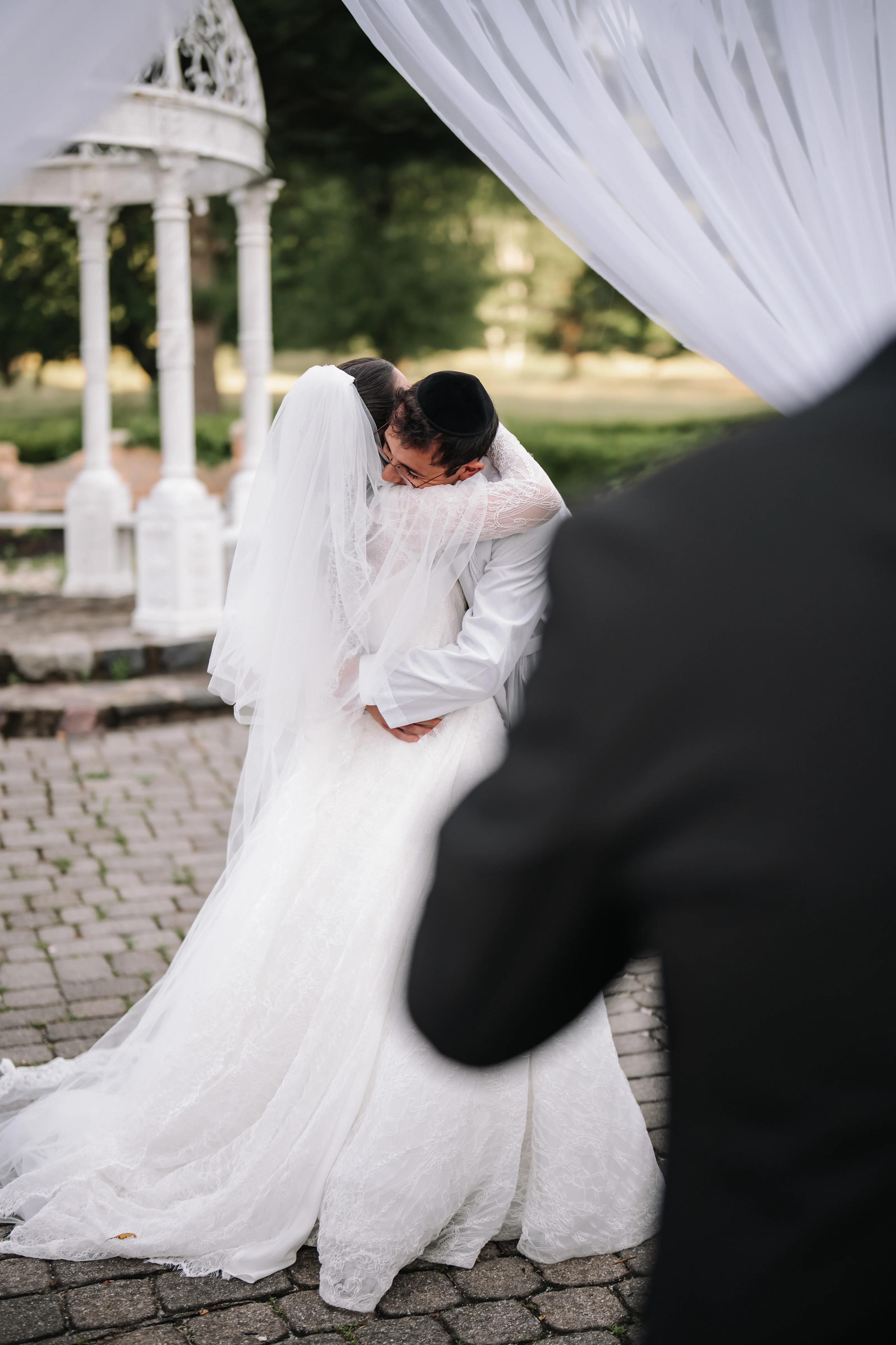 Groom bowing his head in a tender moment as a bridal veil drapes across the scene outdoors near white columns.