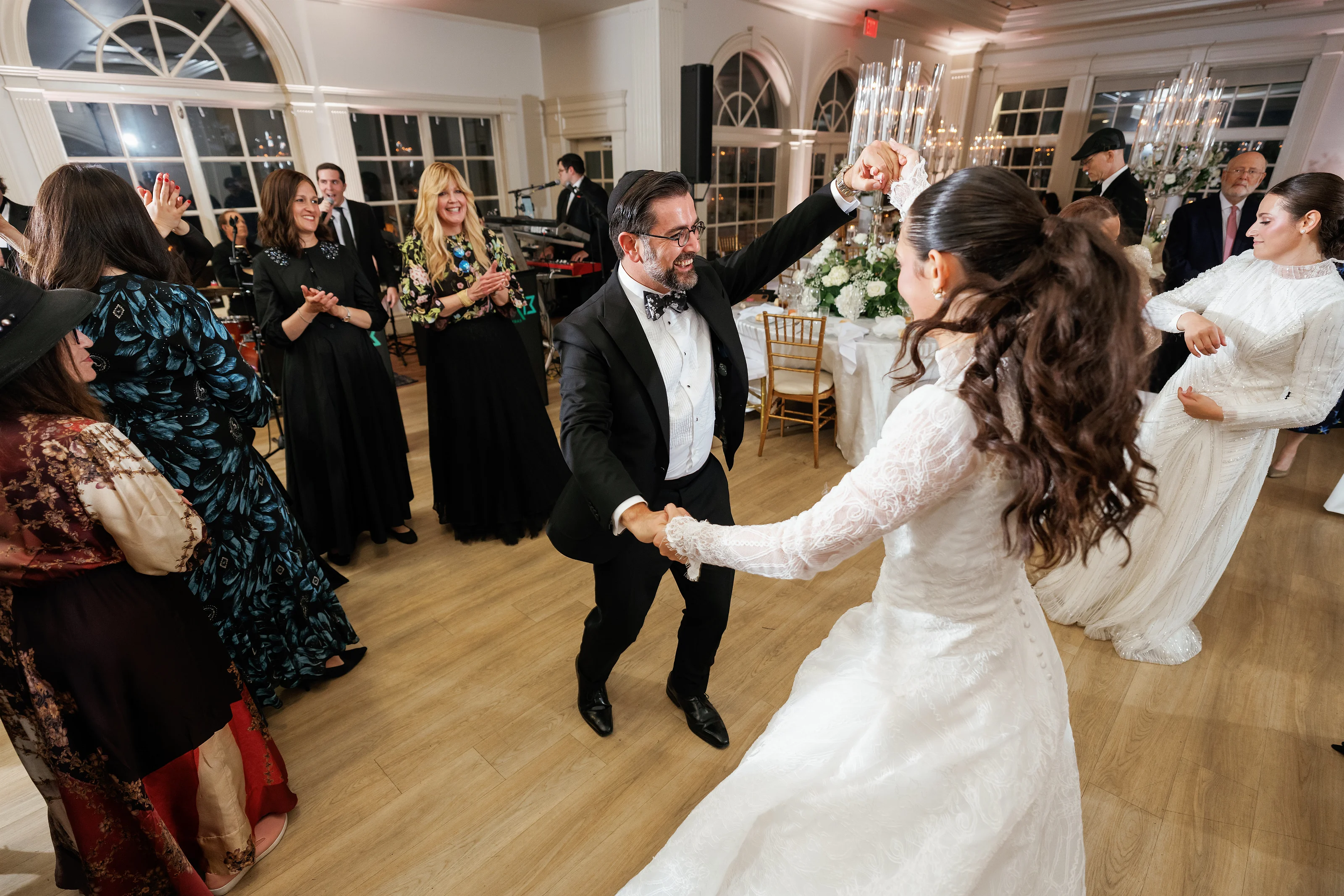 Groom and bride dancing together on the reception floor while guests clap and cheer in a warmly lit ballroom.