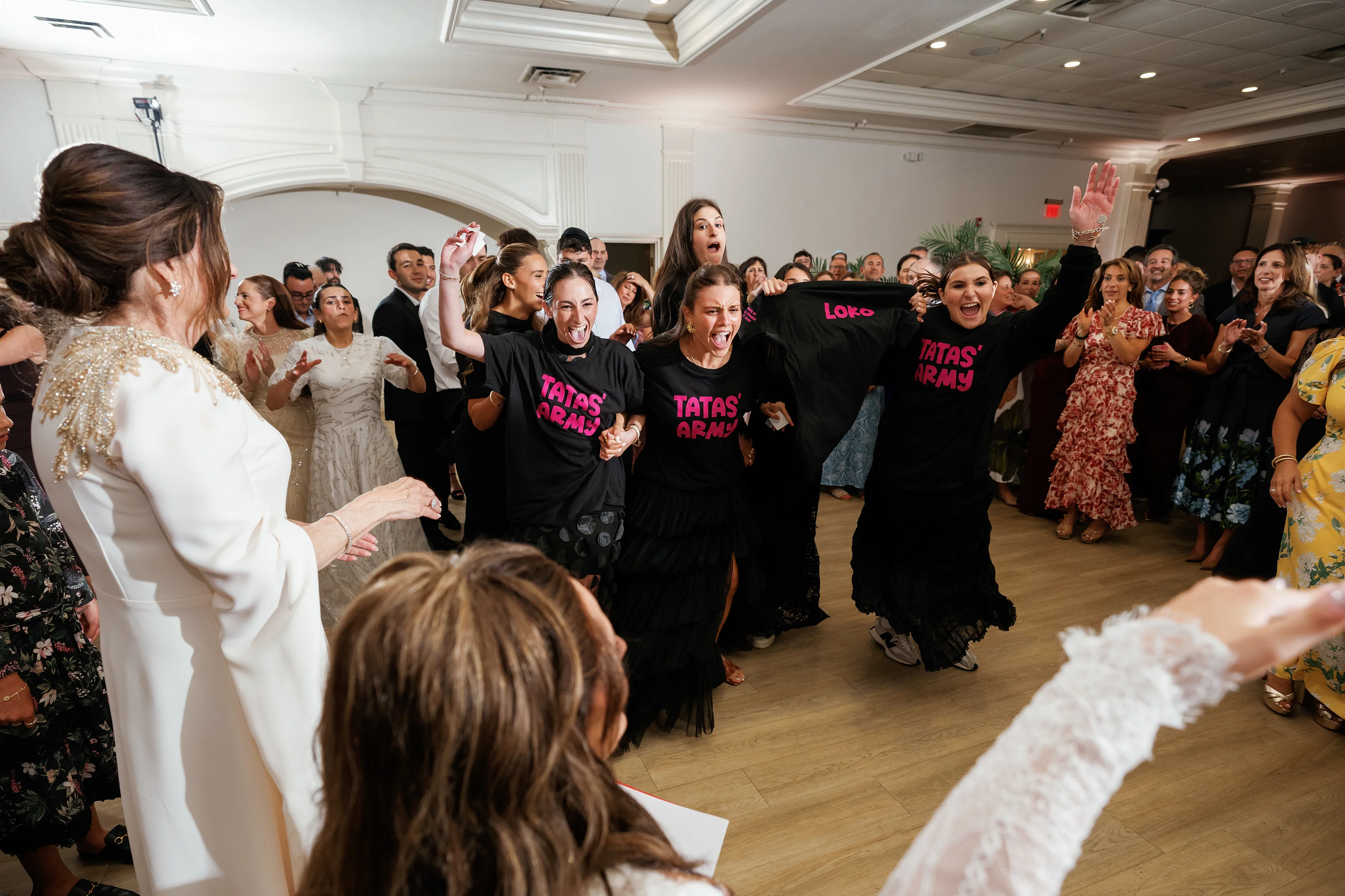 Candid wedding dance moment in an indoor ballroom with the bride in the foreground and guests in black shirts laughing and cheering with raised hands.