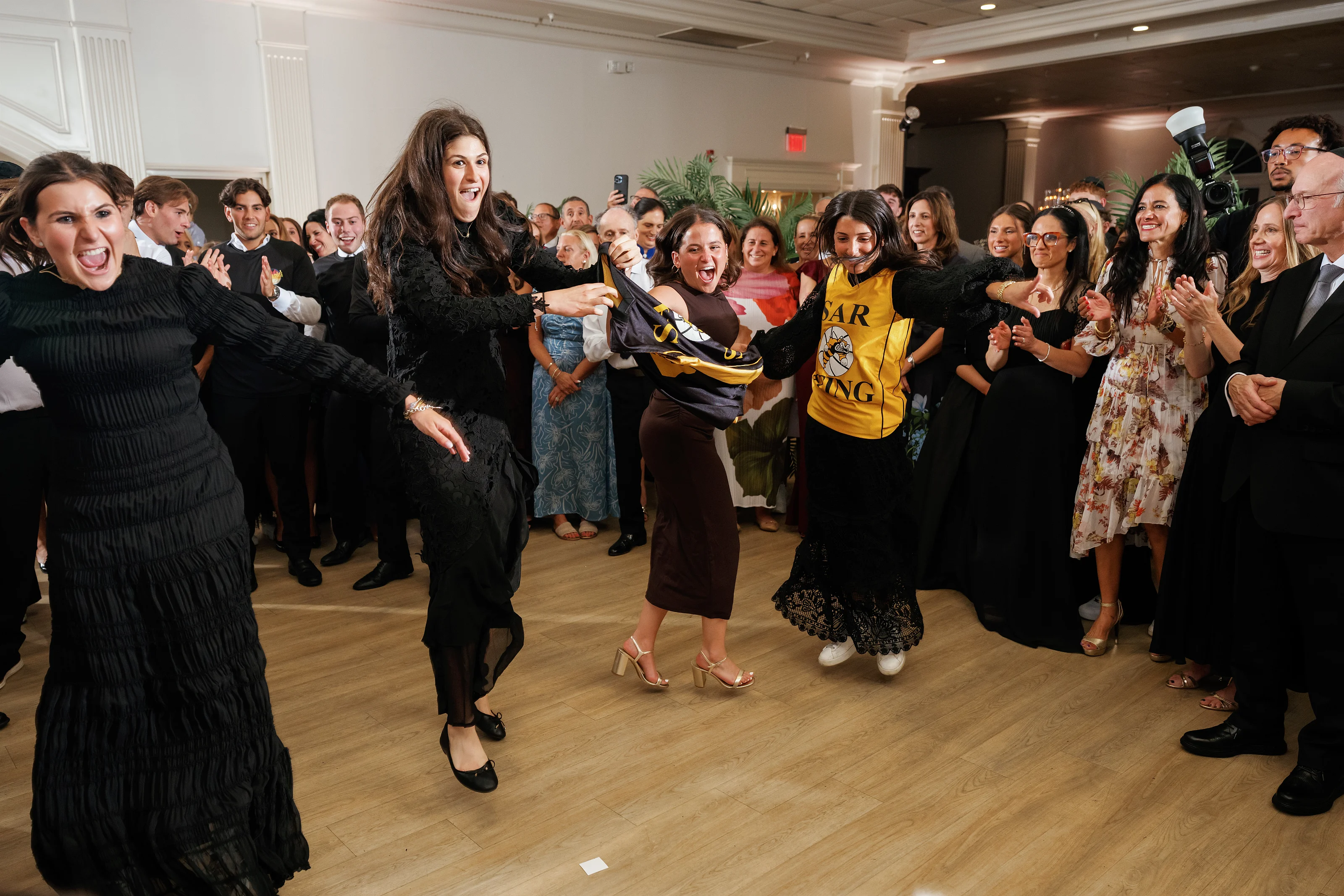 Celebratory dance-floor moment in a ballroom, with guests clapping and the central bride and groom figures laughing mid-celebration.