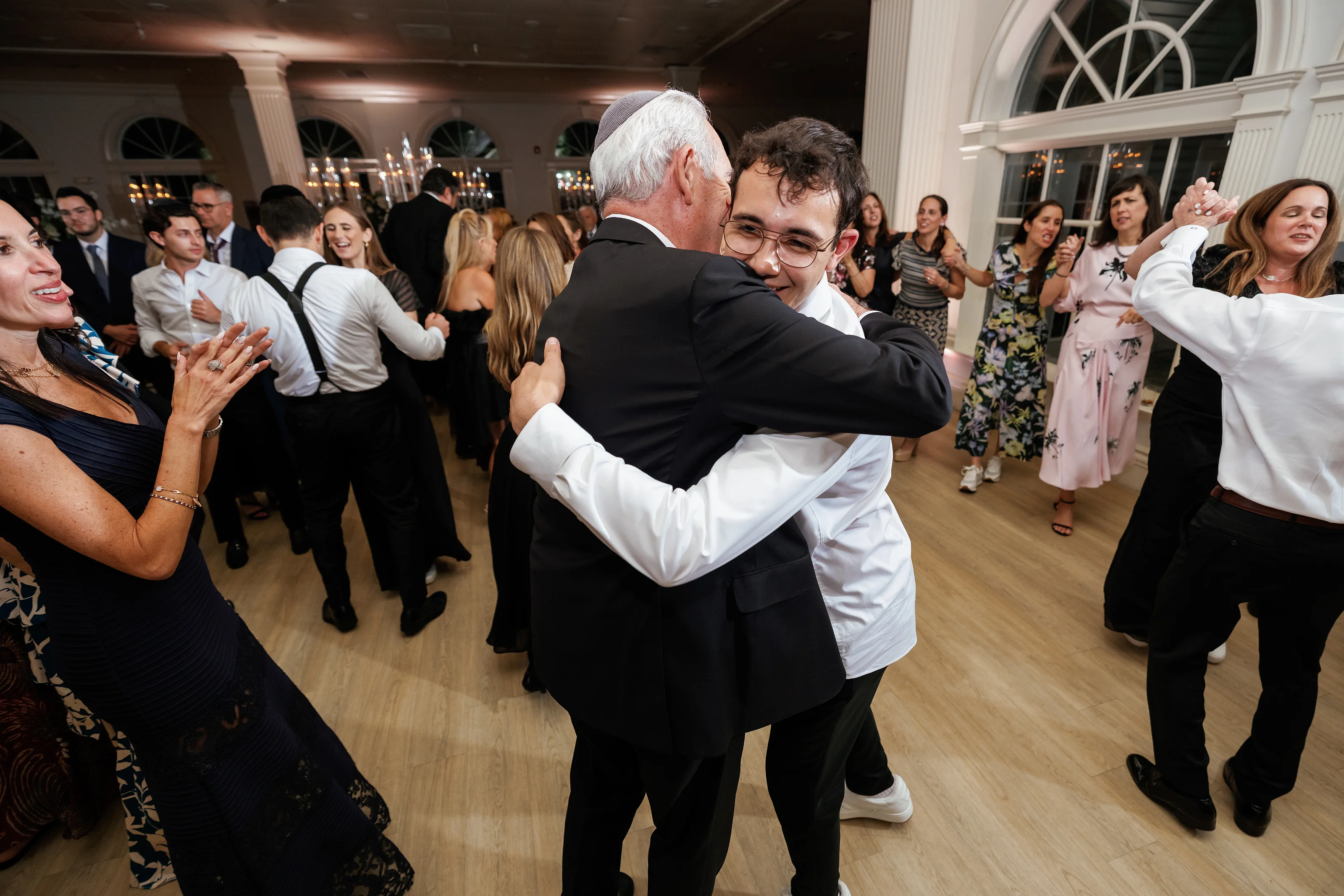 Couple embracing mid-dance at a celebration, with guests clapping and smiling in a warmly lit hall.