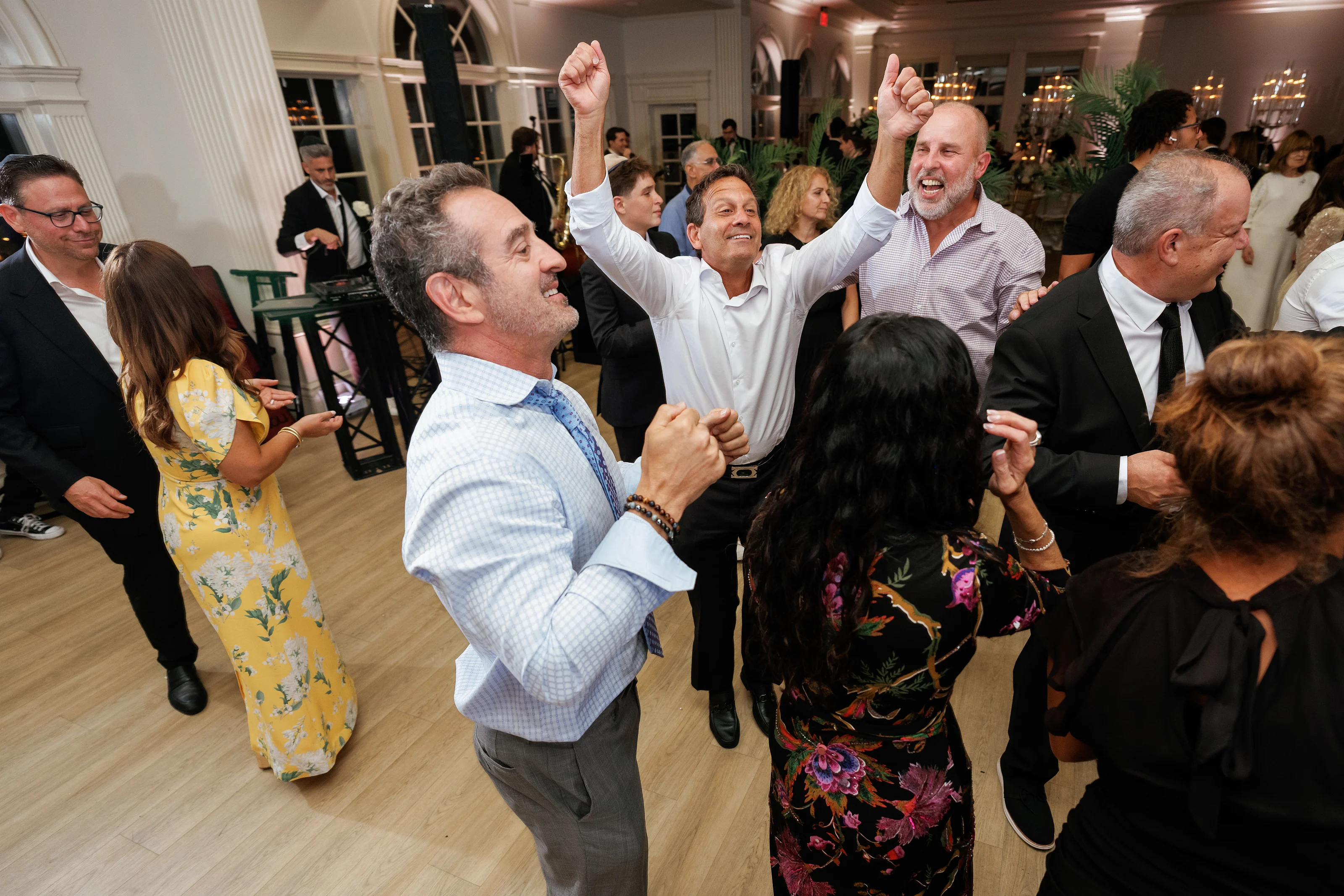 Candid dance-floor moment with multiple guests smiling and raising their arms during a celebration in an indoor reception hall.