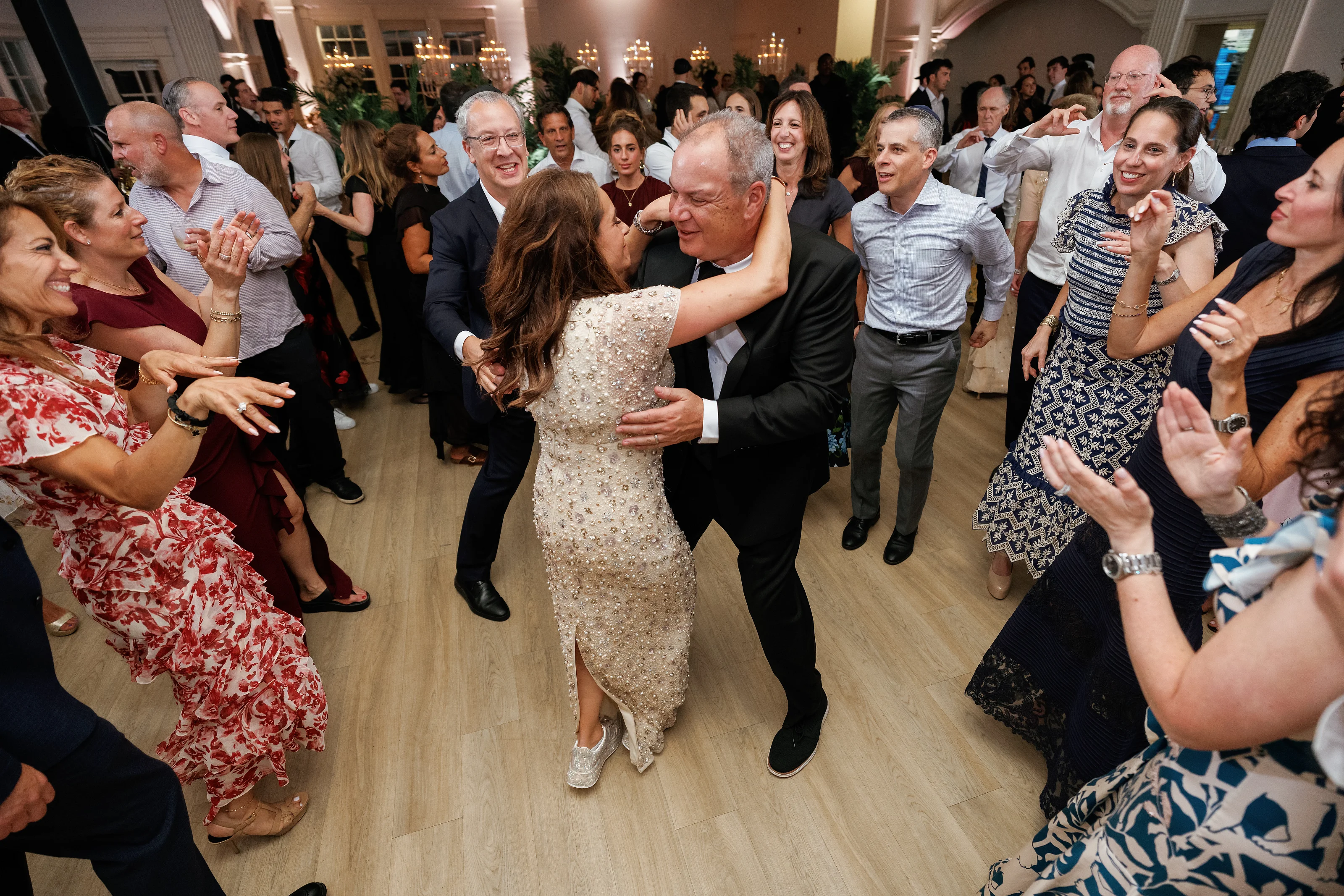 Couple dancing at a wedding reception surrounded by guests clapping, with warm indoor lighting and smiling faces.