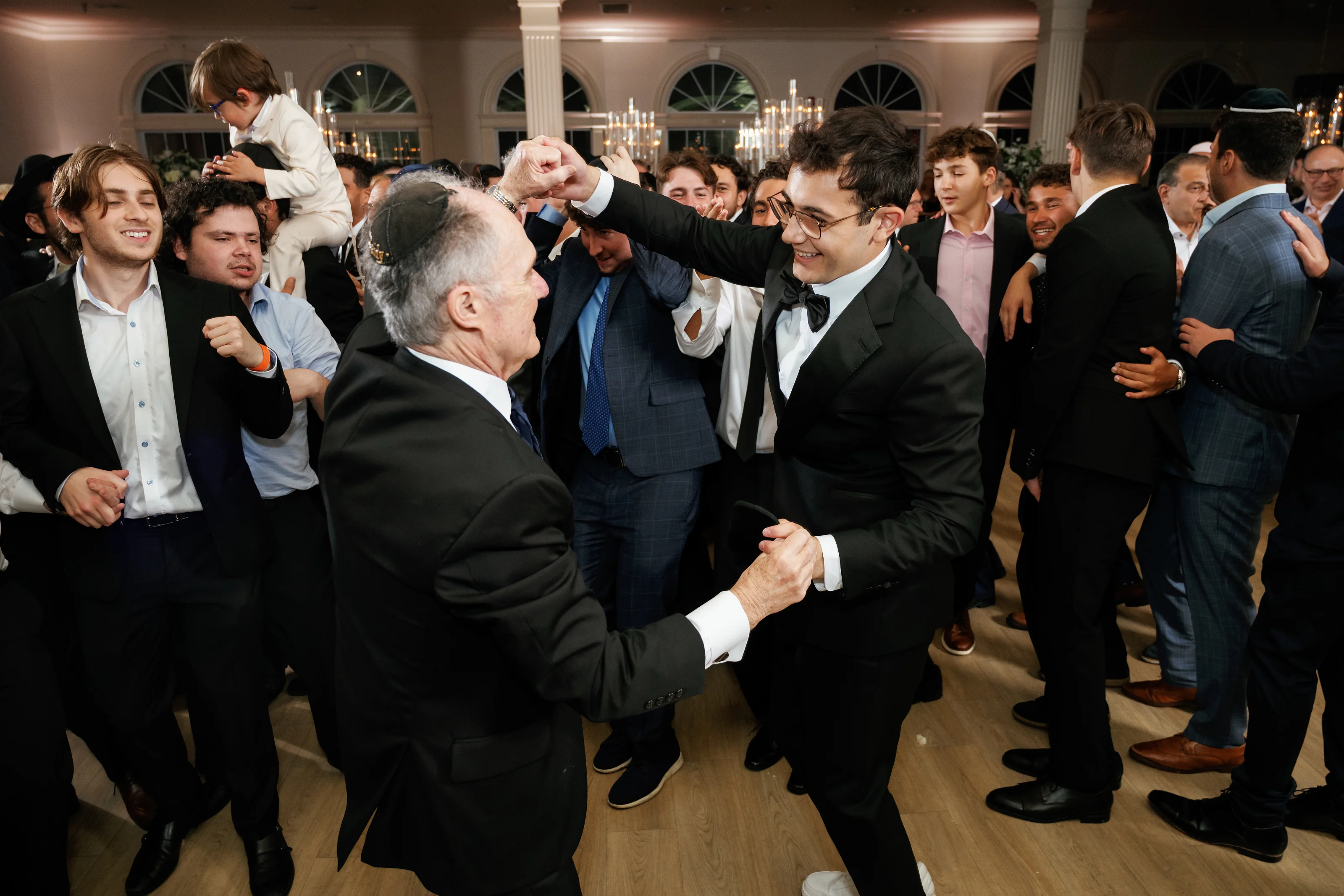 Jewish wedding reception dance-floor moment with a smiling man in a tuxedo surrounded by friends and family in formal attire, warm indoor lighting, and lively celebration.