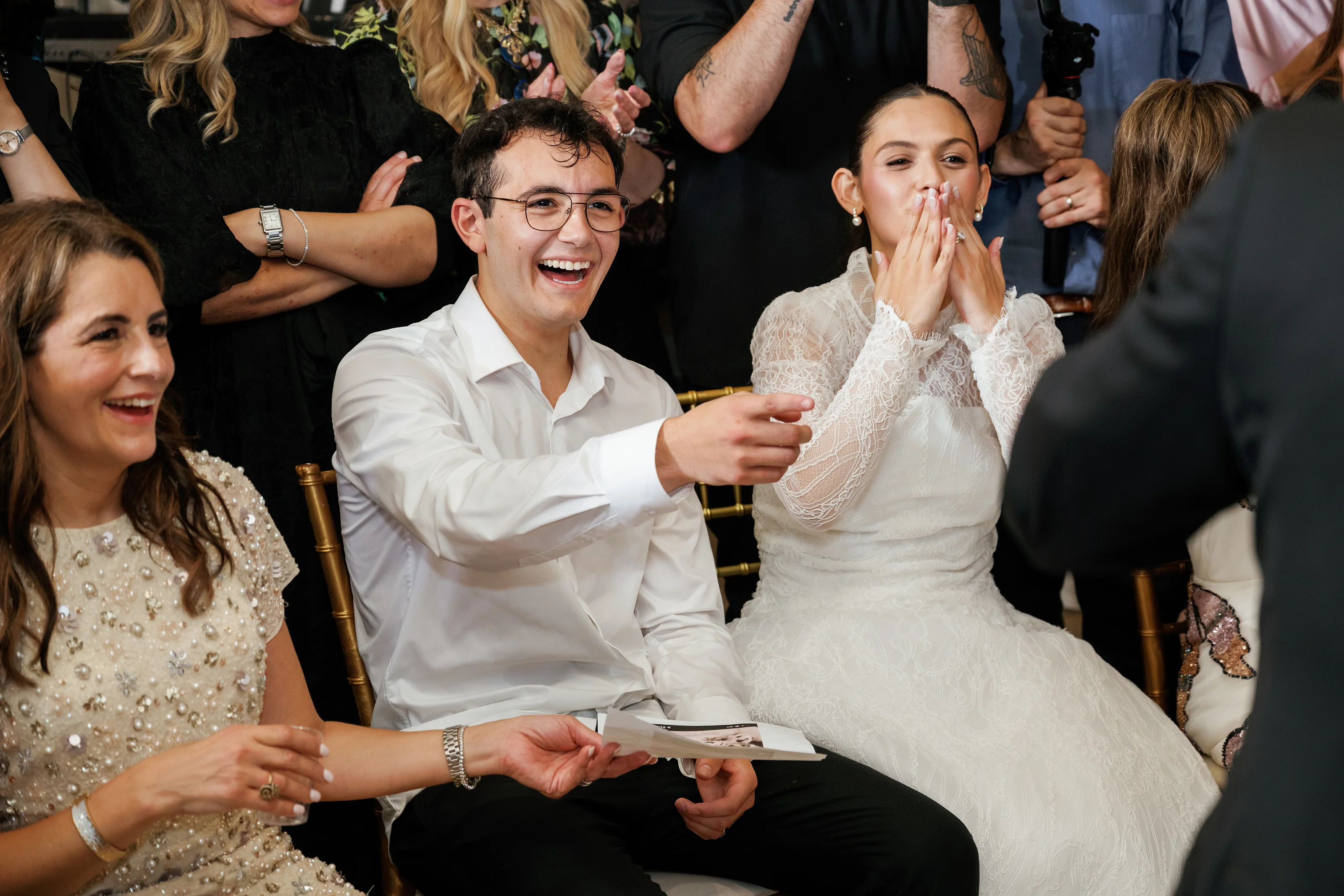 Smiling bride covering her mouth with her hands while the groom laughs beside her during a joyful wedding celebration with close family seated around.