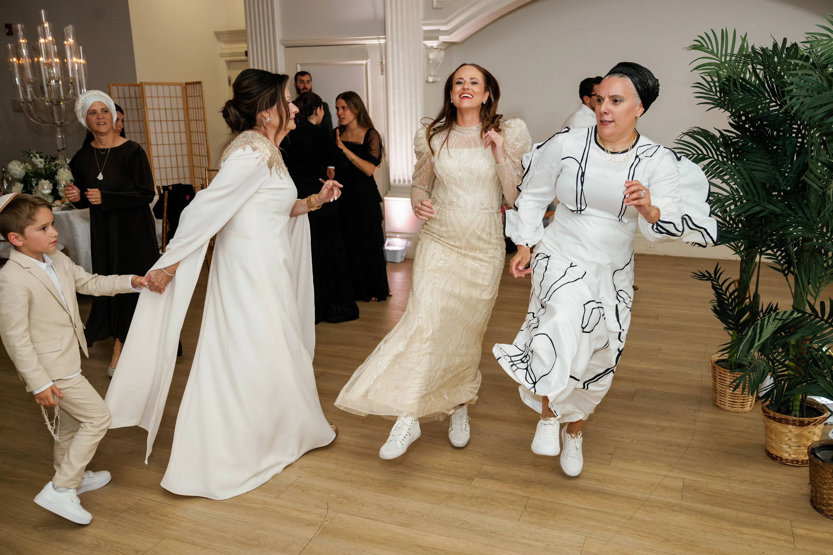 Bride smiling while walking forward mid-dance with guests in traditional attire at an indoor celebration.