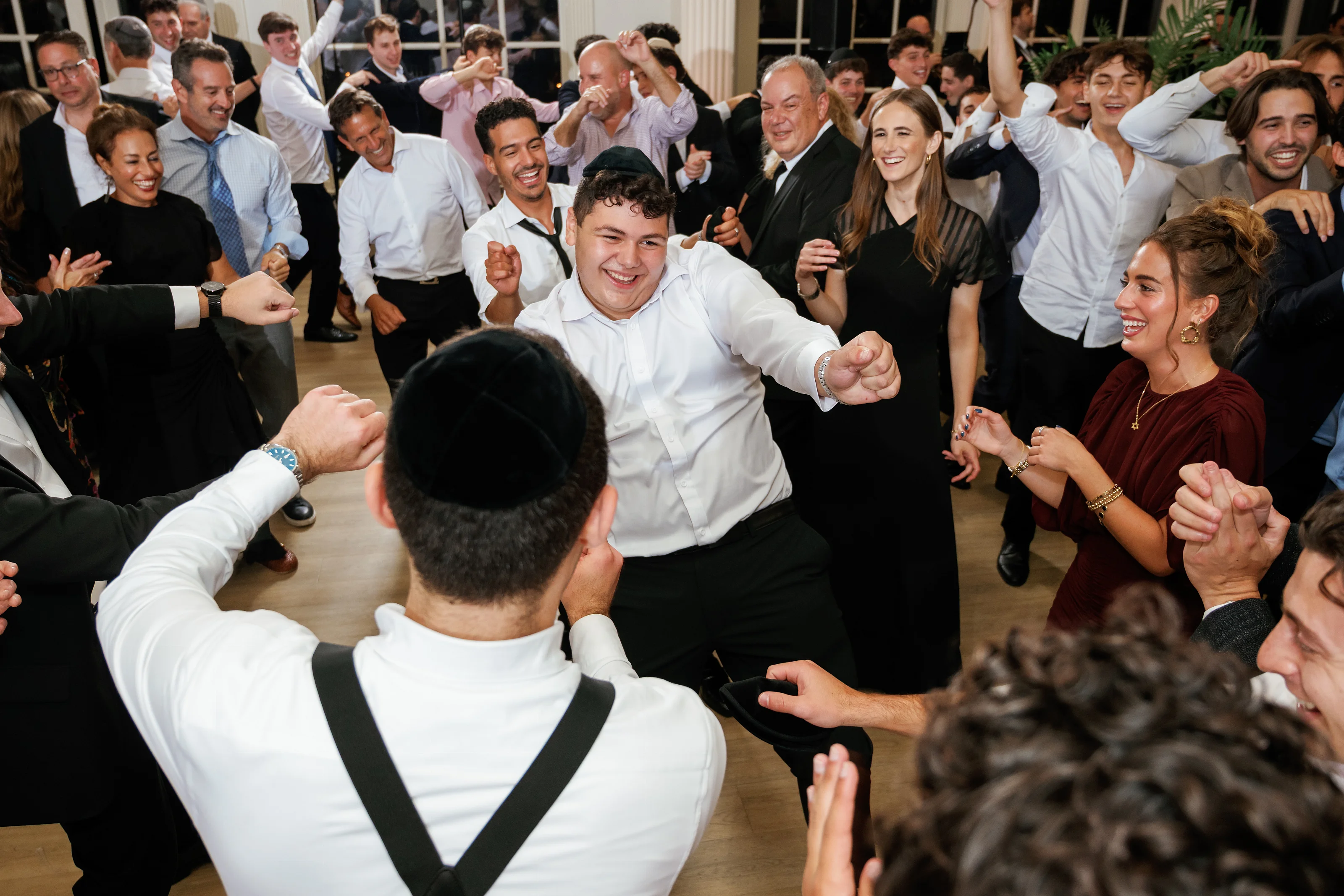 Hora dance moment at a wedding with the groom smiling in the foreground while guests clap and raise their arms.