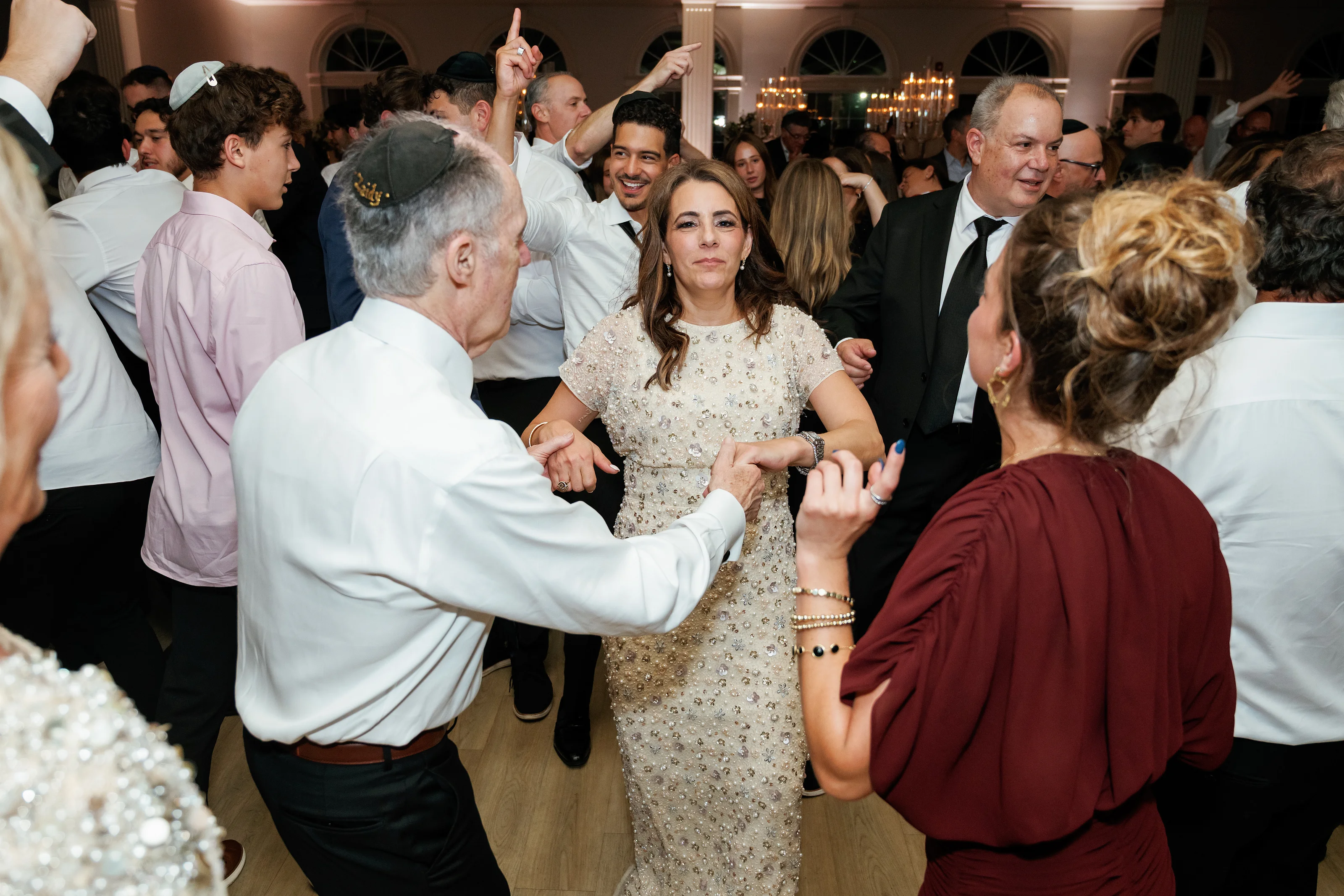 Bride centered in the middle of a lively wedding dance circle, smiling as guests surround her indoors under warm chandeliers.