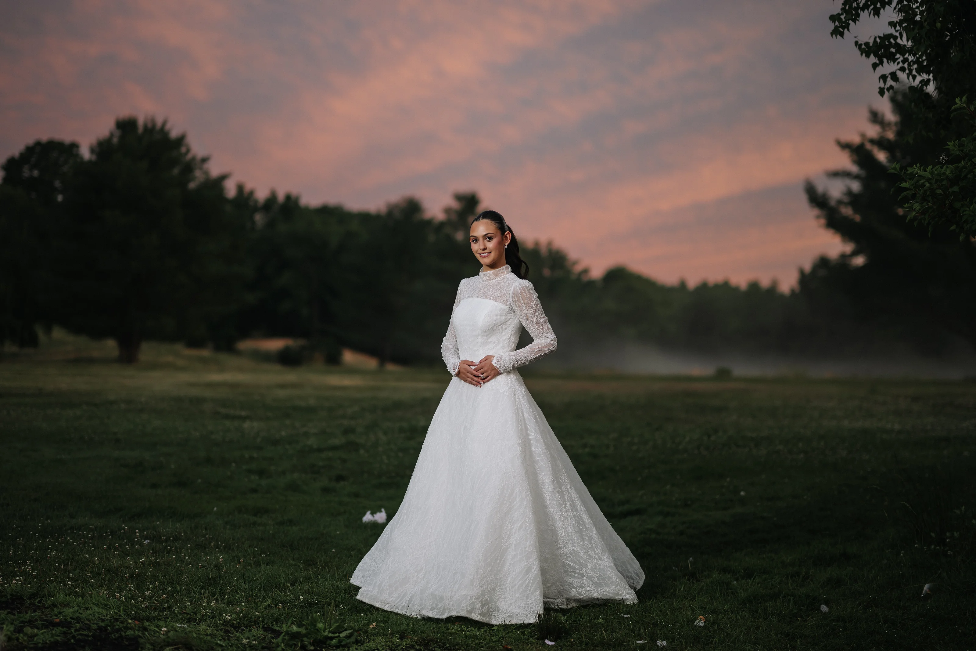 Bride in a white wedding gown posed on a grassy field at sunset with a pink-orange sky behind her.